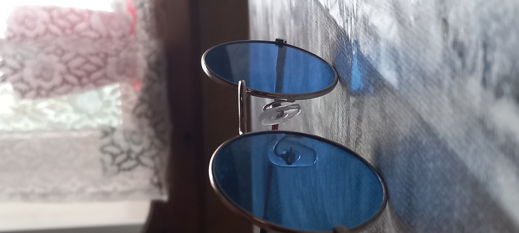 The image presents a close-up, static shot dominated by a pair of blue-tinted sunglasses, positioned vertically with one lens slightly above the other. The sunglasses are in sharp focus, highlighting their round or oval lenses, metallic frame, and clear plastic nose pads.
To the left, the background is softly blurred, revealing a light-colored, possibly white or cream, patterned fabric. This fabric, likely a curtain or decorative textile, features a repeating floral or damask design, suggesting an indoor setting.
On the right side of the frame, a textured surface, which could be a wall or a wooden panel, is visible. This surface displays subtle vertical lines and variations in tone. Notably, there are faint blue reflections cast upon this surface, originating from the sunglasses' lenses, along with patterns of light and shadow.
The lighting appears to be natural, likely daylight, entering from the left side of the frame. This creates a sense of depth and accentuates the reflective qualities of the lenses. There are no people or discernible activities taking place. No text is visible within the image. The scene conveys a quiet, detailed still life, set indoors, consistent with a domestic environment in Kryvyi Rih, Ukraine.