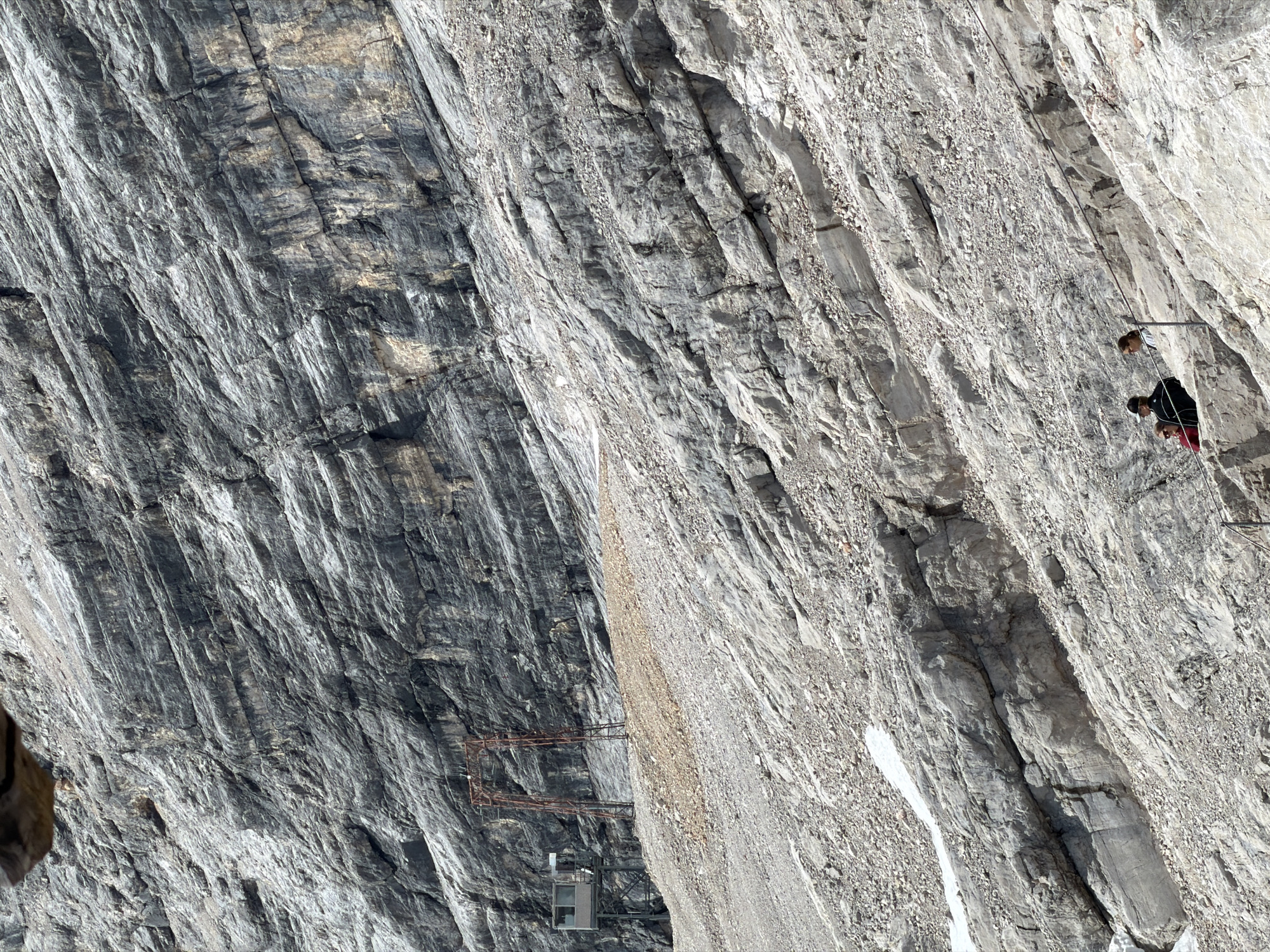 The image depicts a steep, rocky mountainside, likely a section of the Zugspitze massif near Garmisch-Partenkirchen, Germany, given the presence of ski lift infrastructure. The terrain is predominantly gray rock, with layers of scree and scattered patches of snow, indicative of a high-altitude environment.
In the mid-ground, an industrial structure with a cabin-like building and a tall, rusted metal pylon stands against the rock face. This is likely part of a cable car or ski lift system.
Further down the slope, in the lower right quadrant, three individuals are visible. One person, wearing a dark jacket and a cap, is seated. Another person, wearing a red top, is standing behind the seated individual. A third person, with long brown hair, stands to the right of the seated person, leaning on a railing. They appear to be observing the view or resting. A wire rope runs horizontally in front of them, part of a safety barrier or a different lift system. The people are positioned on a rocky ledge with a steep drop behind them. The overall scene conveys a sense of a rugged, mountainous landscape with human activity integrated through the infrastructure.