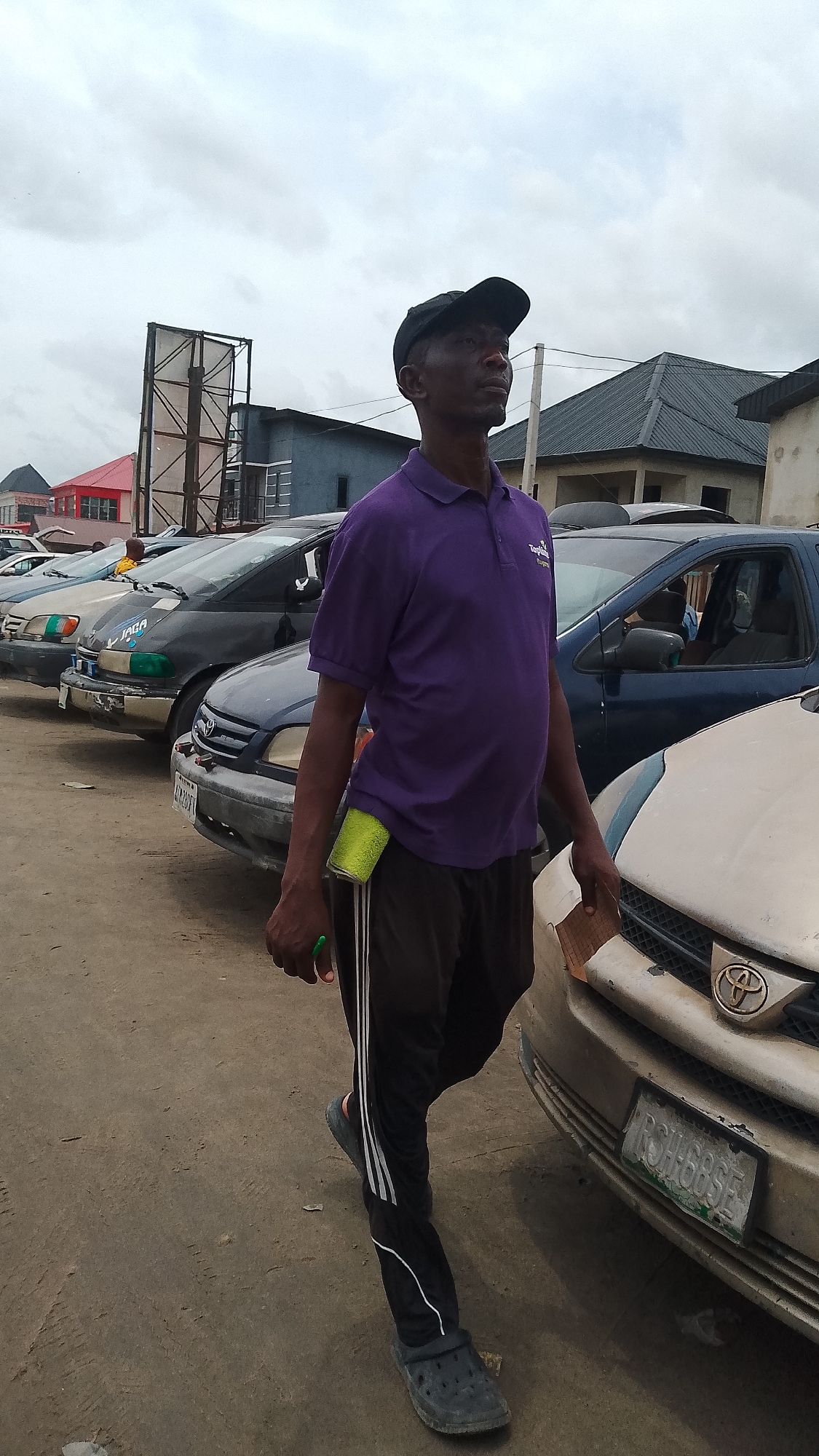 An adult Black man is walking in a parking lot located in Unknown, Unknown. He is wearing a purple polo shirt with a logo on the chest, black track pants with white stripes down the sides, and dark-colored Crocs. He has a black cap on his head, and he is holding a piece of paper in one hand and a pen in the other. A green cleaning rag is tucked into his pants. Several cars are parked in a row behind him, and the background includes buildings and an overcast sky.