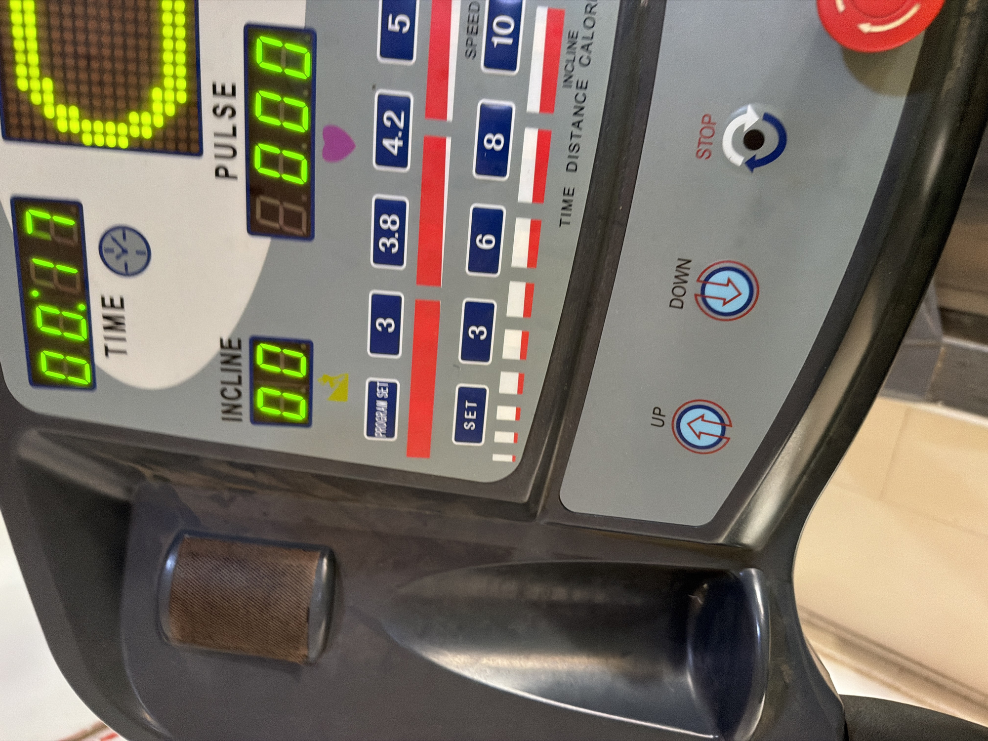 The image is a close-up shot of the console of a dark gray treadmill. The console features a digital display with green illuminated numbers. The top display shows "00.77" under the word "TIME" with a small clock icon. To the right of the time display, a segmented LED display shows a green "C" shape. Below the time display, "INCLINE" is labeled, with a display showing "0.0". To the right of the incline display, "PULSE" is labeled, with a display showing "8.000". There is a small pink heart icon below the pulse display.
Below these main displays are rows of buttons. The top row of buttons are blue and labeled "PROGRAM SET", "3", "3.8", "4.2", and "5". Below this row are red horizontal bars. The second row of blue buttons are labeled "SET", "3", "6", "8", and "10". Below this row are alternating red and white horizontal bars. Text at the bottom of this section reads "TIME DISTANCE INCLINE CALOR".
Further down the console are two circular buttons with arrows. The left button is labeled "UP" and has an upward-pointing arrow. The right button is labeled "DOWN" and has a downward-pointing arrow. To the right of these is a circular button labeled "STOP" with a circular arrow icon. A large red emergency stop button is visible in the bottom right corner of the image.
To the left of the console, a textured, brown cylindrical object is visible, possibly a cup holder or speaker. The treadmill is made of dark gray plastic, and the console is a lighter gray with black accents.
This image does not contain any information about the city of Gura Top, Nigeria, or any people.