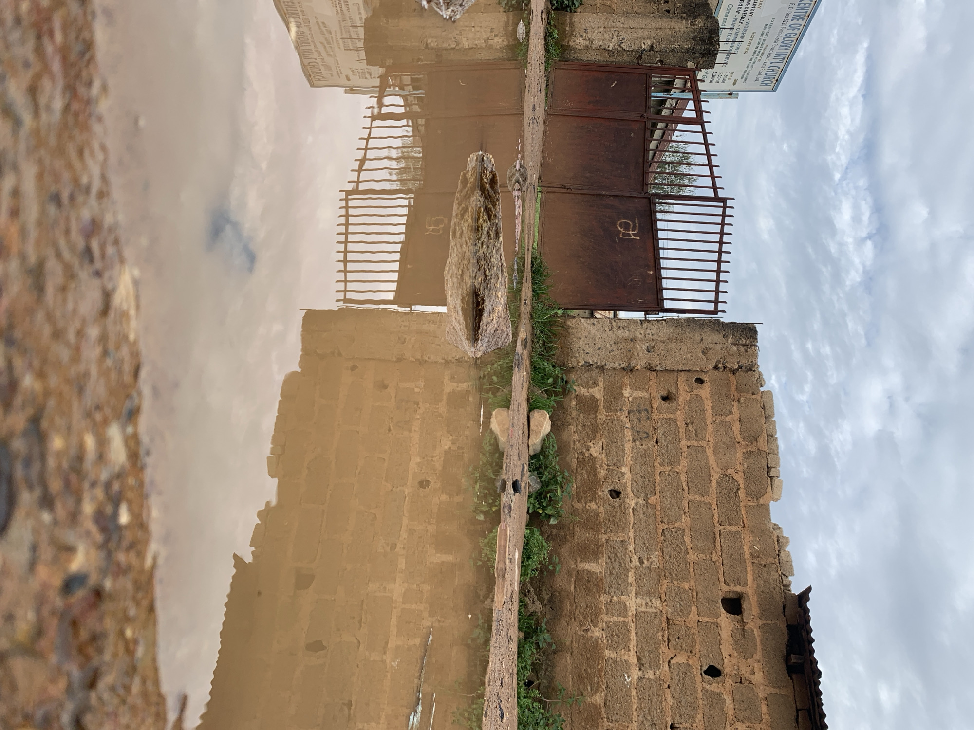 The image depicts an exterior scene in Rafin Par, Nigeria, characterized by a large, murky puddle dominating the foreground and midground. The puddle, filled with reddish-brown water, reflects the overcast sky and the structures beyond. In the upper half of the image, a weathered, rusty double-leaf metal gate with vertical bars is visible, set between two concrete pillars. A stylized white symbol is painted on the right gate leaf. To the left of the gate, a blue and white sign for "CENTRE OF GLORY INT'L CHURCH" is partially legible, displaying an address (P.O. Box 5260 Farm-Gada Road, Zaria Road, Jos) and listed activities with times. To the right of the gate, an unplastered wall constructed from reddish-brown concrete blocks extends horizontally; the top edge appears irregular and partially broken, and several dark, circular perforations are present in the blocks. Sparse green vegetation grows along the base of this wall and near the gate pillars. Behind the gate, parts of a structure with a dark, corrugated roof are visible. The immediate foreground consists of muddy, reddish-brown soil mixed with small stones, leading into the puddle. Several rocks are positioned within or at the edge of the puddle, with one prominent elongated dark rock near the center. The sky above is largely cloudy and grey. No people are visible.