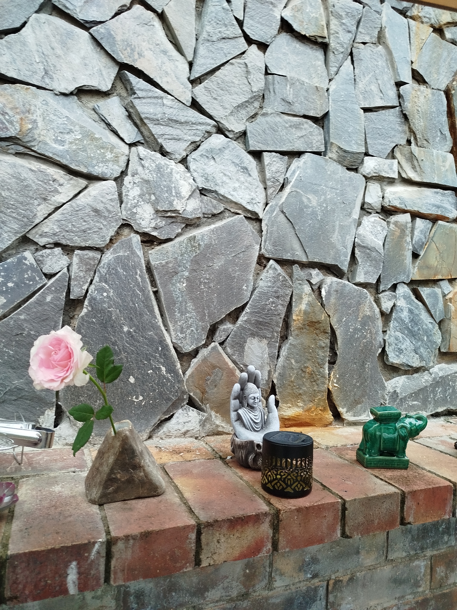 The image shows a low brick wall in front of a stacked stone wall. On the brick wall are several decorative objects: a pink rose in a triangular vase, a statue of a Buddha in cupped hands, a small black cylindrical lantern with ornate gold cutouts, and a green ceramic elephant figurine. The stacked stone wall behind the objects consists of irregularly shaped gray stones. The location is unknown but the user has specified Lạc Dương, Vietnam.