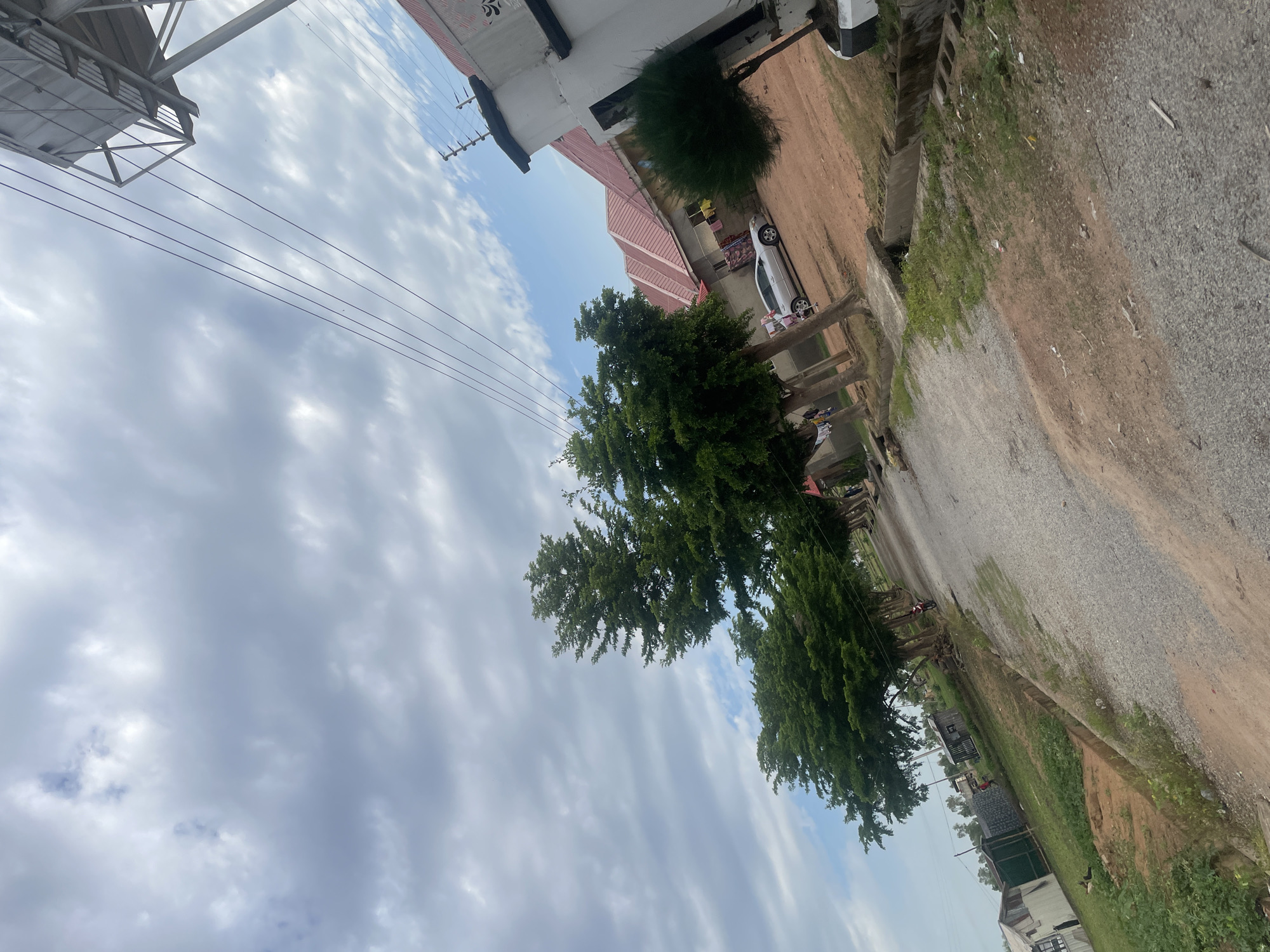 The image depicts a street scene in Jauro Yinu, Nigeria, under a cloudy sky. The perspective is from a low angle, looking upwards.
The scene features several subjects: a white car parked on a dirt road, a building with a red roof and white walls, and several lush green trees. There are also power lines crisscrossing the sky, supported by poles. In the foreground, a gravel path runs alongside the dirt road, with some sparse vegetation and debris. The background shows more of the road and some structures.
No specific activity or event is clearly taking place, though the presence of the car and buildings suggests a residential or commercial area.
Notable details include the overcast weather, indicated by the cloudy sky. The time of day appears to be daytime, likely late morning or afternoon given the lighting. The environment looks somewhat unkempt, with visible debris along the roadside. There is no visible text in the image.