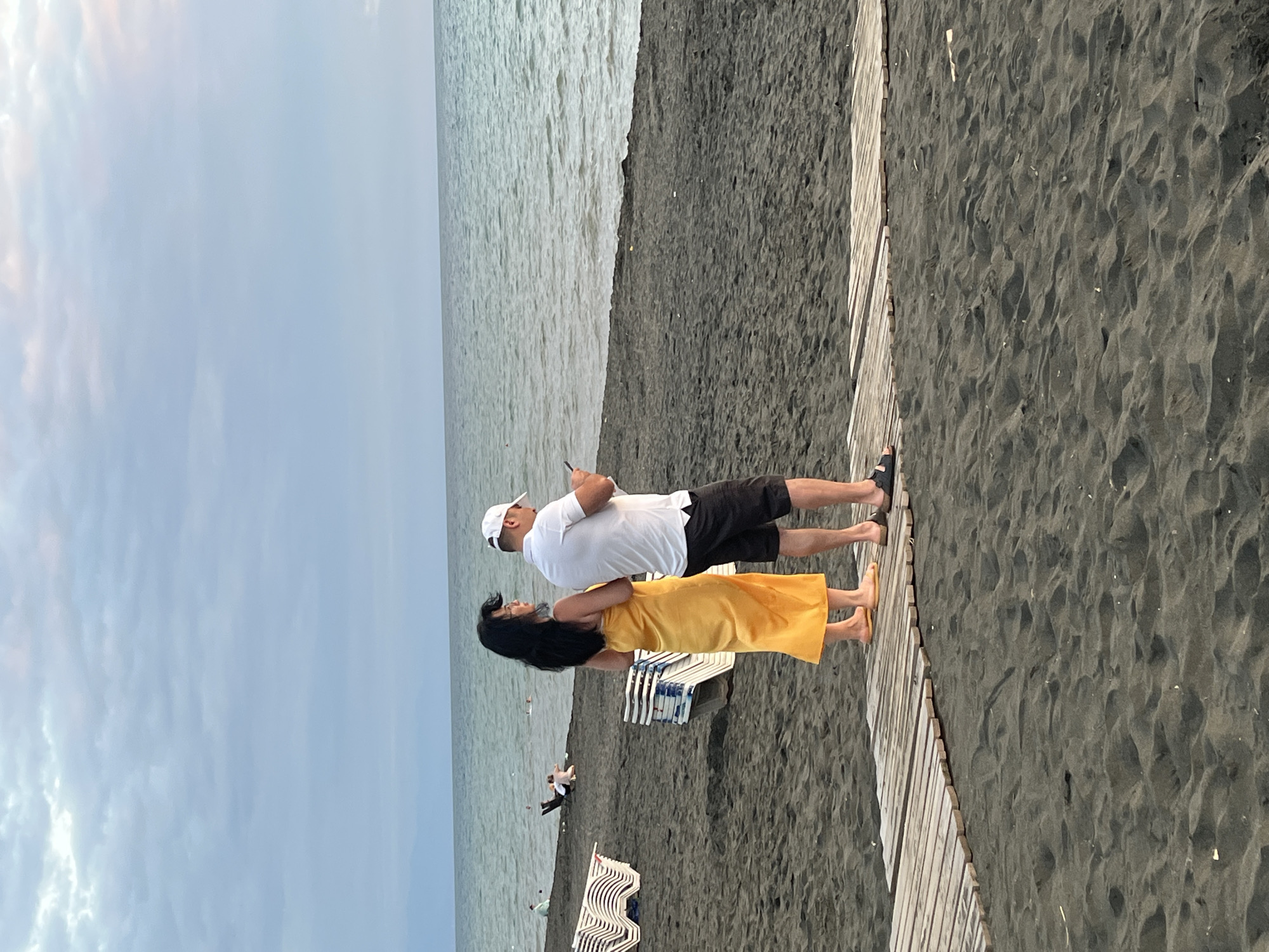 The image depicts a beach scene in Ureki, Georgia, with a focus on two individuals standing on a wooden walkway. A woman wearing a bright yellow wrap dress stands on the left, and a man in a white polo shirt and black shorts is on the right. The man is holding a mobile phone and appears to be looking at it. The beach has dark, coarse sand. In the background, the sea is visible with gentle waves under a cloudy sky. Several rows of white beach chairs are stacked on the sand to the left of the walkway. Further in the distance, on the beach and in the water, other people are present, including some individuals sitting on chairs or lying down on the sand, and some swimming in the sea. The overall impression is a moderately populated beach during what appears to be late afternoon or early evening.