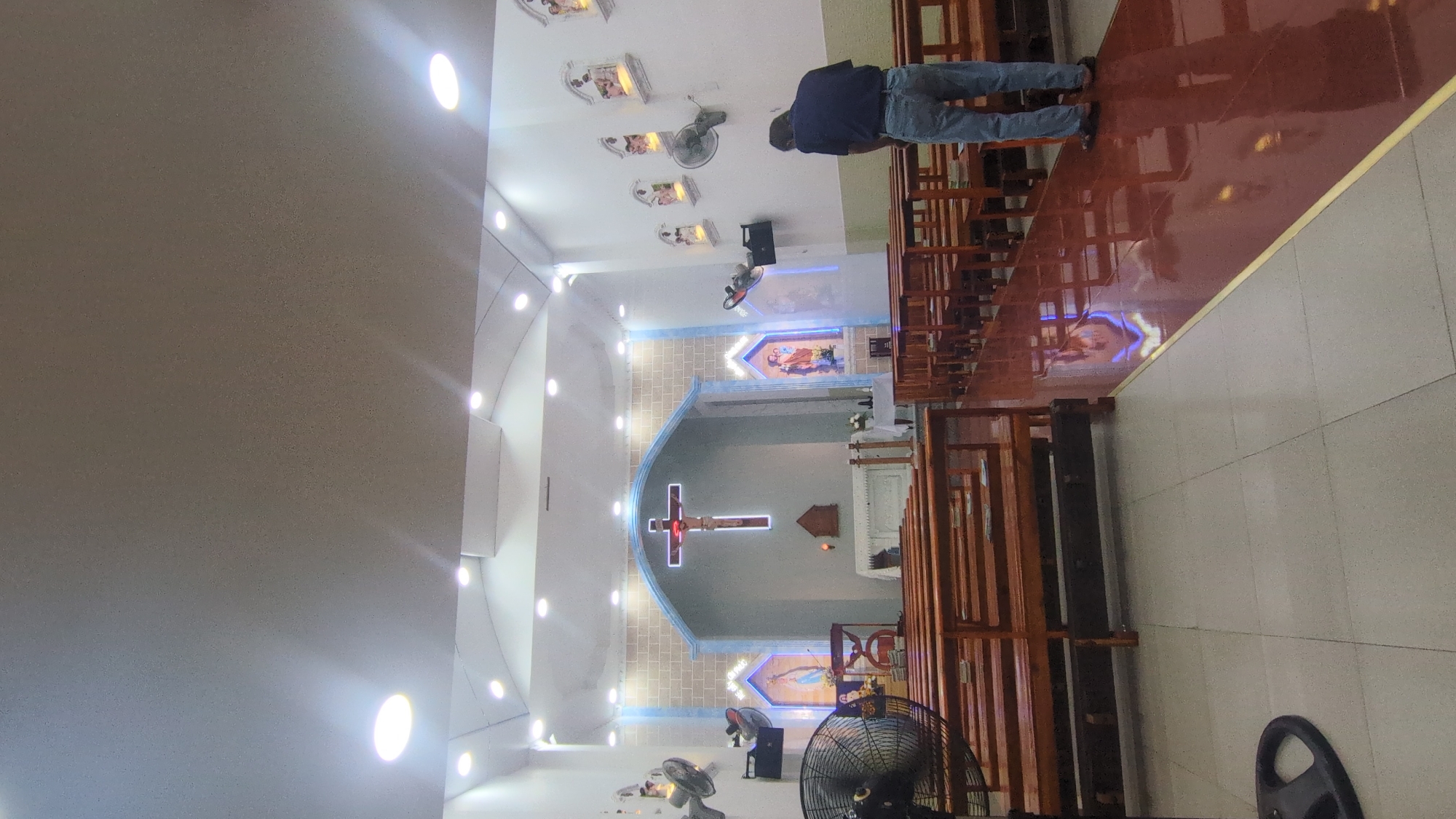 This image, oriented vertically but described as if correctly rotated, captures the interior of a brightly lit church or religious building, likely in Thủ Đức, Vietnam.
The main subject is the church interior itself, with a person standing on the right side, facing towards the altar. This individual is dressed in blue jeans and a dark top, appearing to observe or pray. On the right, several rows of traditional wooden pews are visible.
The focal point of the church is the altar area, featuring a large crucifix with Jesus on the cross, strikingly illuminated by blue and white neon lights. Above the crucifix, within a semi-circular archway detailed with light brick-like textures, a religious statue is placed in a lit niche. Another similar niche with a statue is seen to the right of the altar. Along the upper parts of the walls, particularly on the right, several smaller, individually lit shrines or displays, possibly depicting the Stations of the Cross, are mounted.
The church is well-illuminated by numerous recessed lights embedded in the white ceiling and along the architectural curves of the walls. Multiple ceiling fans and a large standing fan in the foreground suggest a warm climate. The floor is divided into two sections: a highly polished, reddish-brown tiled area under the pews, reflecting the altar lights, and a lighter, off-white tiled section to the left, separated by a narrow gold strip.
The scene conveys an atmosphere of peace and solemnity, indicative of a place of worship. The lone person suggests a moment of individual prayer or reflection rather than a public service. No discernible text is visible in the image.