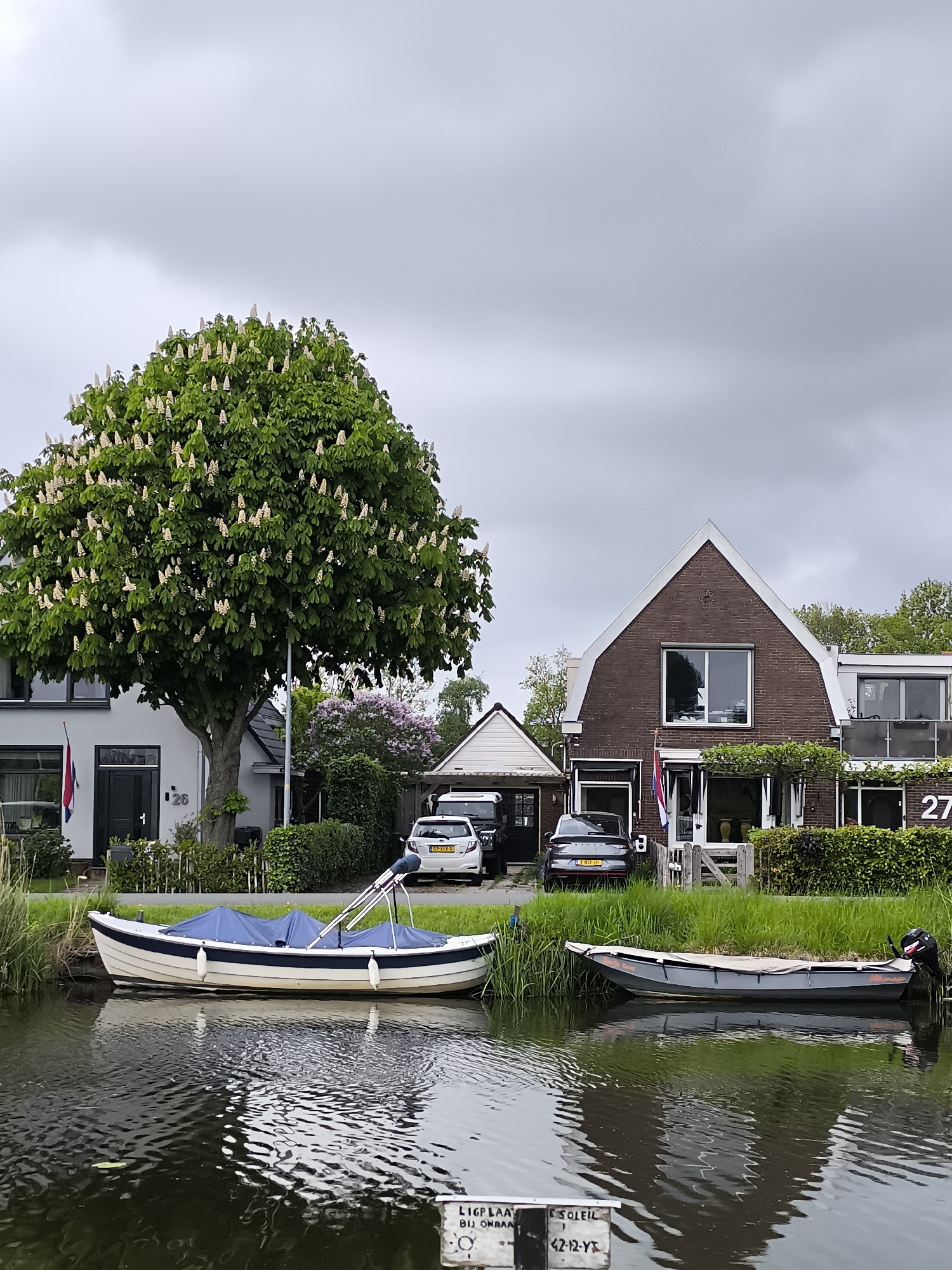 The photograph depicts a canal scene in Oudorp, Netherlands, under an overcast sky. Two boats are moored along the canal bank. The first boat, positioned in the left foreground, is white with a navy blue trim and a blue cover. The second boat, on the right, is gray with an outboard motor attached at the stern. Both are reflected in the water.
Behind the boats, the scene transitions to the land with manicured grass and residential buildings. A large tree with white blossoms dominates the left side of the frame, partially obscuring a light-colored house labeled with the number "26." To the right of the tree, a lilac bush adds a touch of purple to the green landscape. A white car and a dark car are parked in the driveway.
To the right of the cars, a brick house with a steep gable roof is visible, labeled with the number "27." The house has a prominent square window on the second story and greenery surrounding the lower level. A wooden gate is partially visible in front of the house. A Netherlands flag is flown from the front of the property.
In the water in the foreground, a partially submerged signpost is visible, displaying writing, including the word "SOLEIL" and the license plate "42-12-YT."