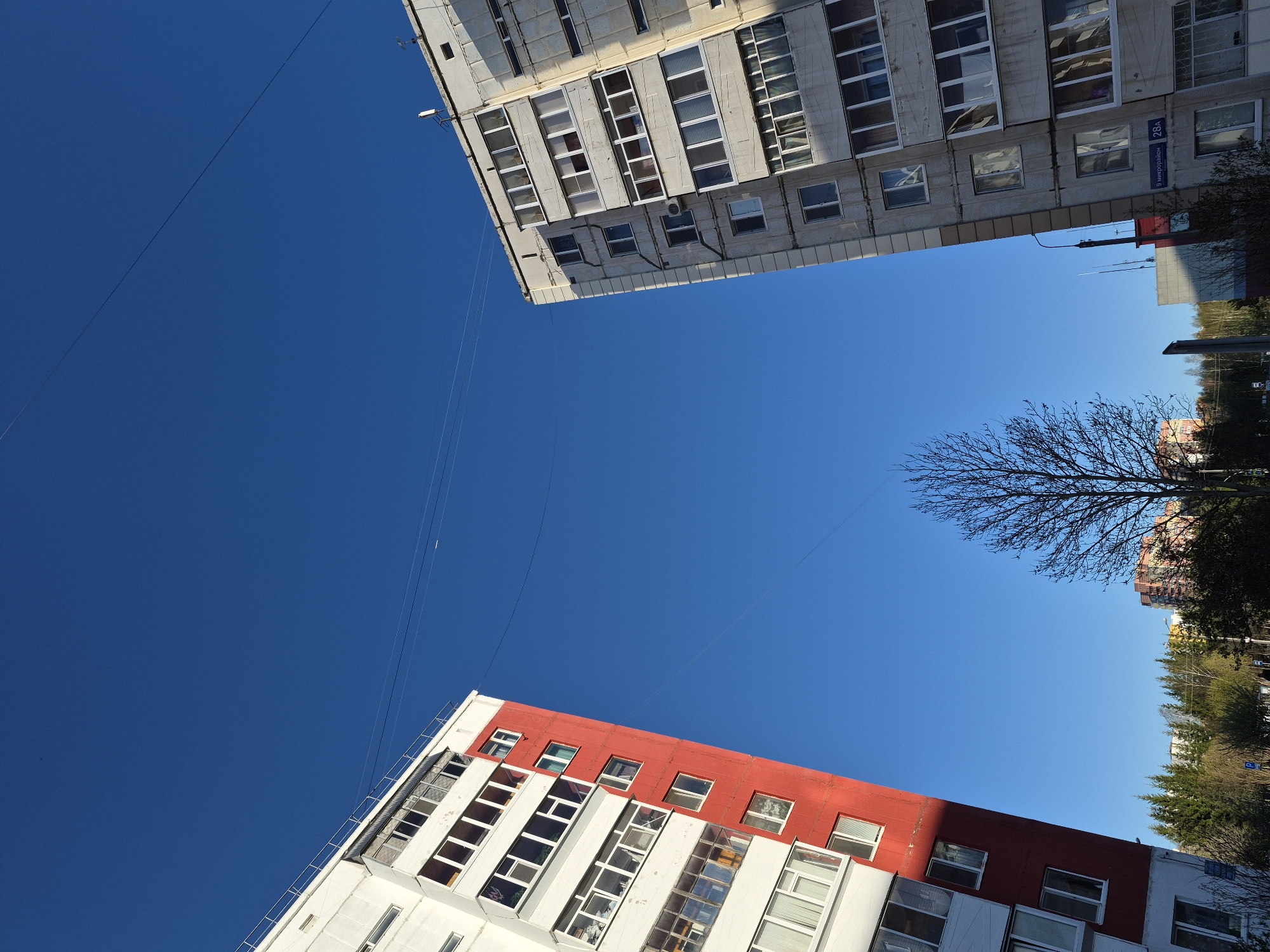 An upward shot reveals two buildings against a bright blue sky. The building on the top right appears to be an older apartment structure with faded walls and rows of windows. A street sign labeled "9 mkr Rayon 28a" is visible on the corner. The building on the bottom left is more modern, featuring a striking red top section above white walls and large window rows. Power lines run across the open blue sky between the buildings. Trees are seen partially in the background on the right. The location is Tobolsk, Russia.