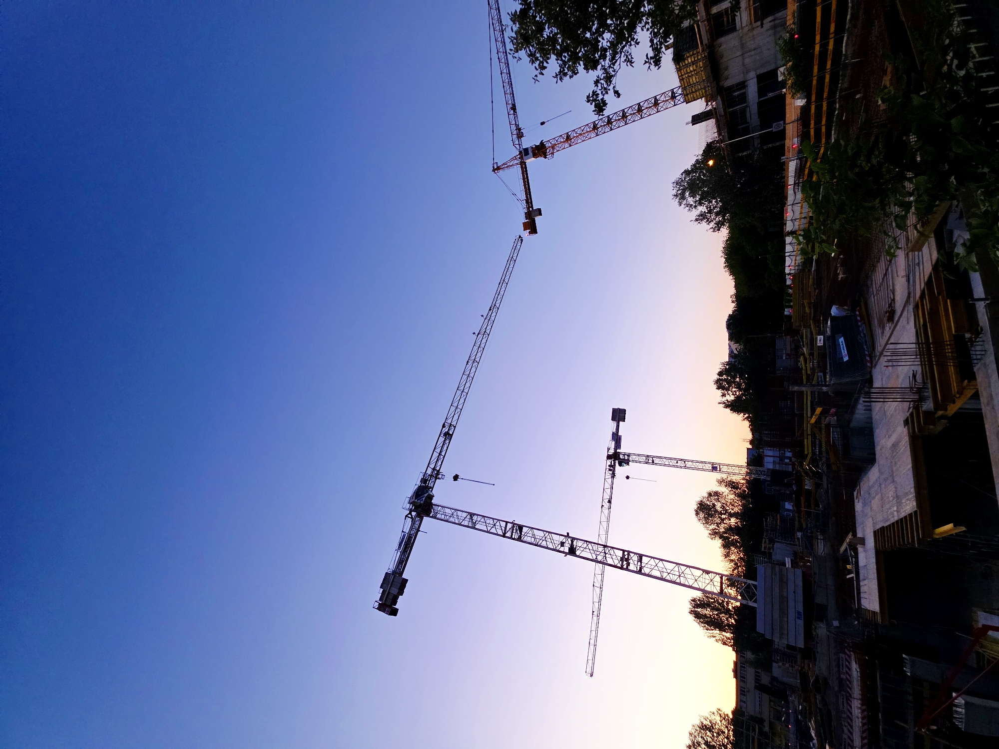This is a photograph of a construction site in Warsaw, Poland, taken at dusk. The sky is a gradient of deep blue at the top, transitioning to a pale purple and then a light yellow towards the horizon. Three large tower cranes are visible against the sky. One crane, on the left, is tall and slender, with its boom extended horizontally. In the center, a second, slightly shorter crane also has its boom extended horizontally. To the right, a third crane is angled upwards, with its boom pointing towards the left.
The construction site itself is a series of concrete foundations, rebar structures, and wooden formwork, indicative of ongoing building work. There are piles of construction materials, including stacked concrete slabs and what appear to be bundles of rebar. In the background, trees line the horizon, and some buildings are partially visible. The lighting suggests it is either early morning or late evening, with artificial lights visible in some parts of the site. The overall impression is one of industrial activity under a serene twilight sky.