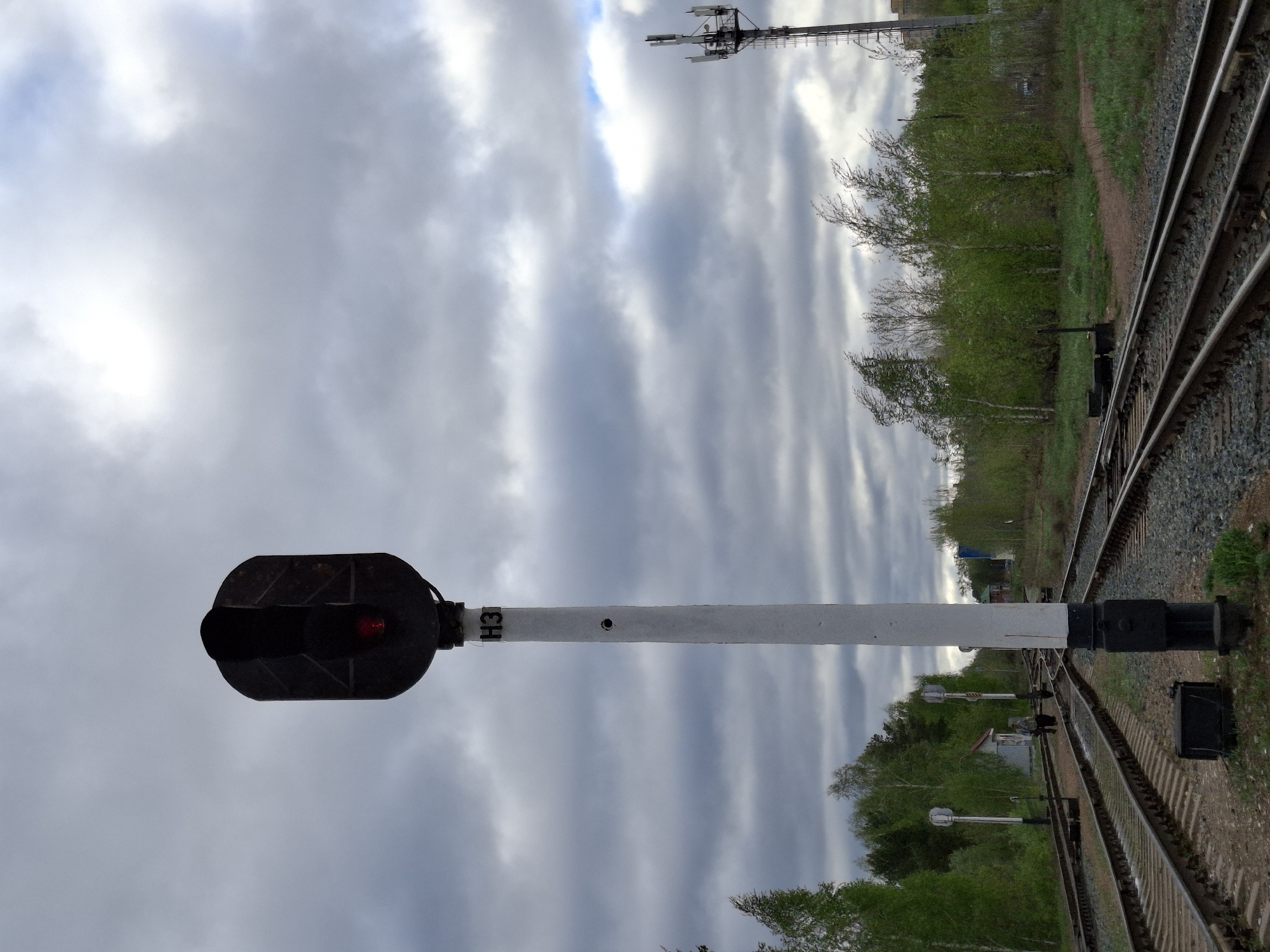 The image depicts a railway scene in what appears to be Chaikovsky, Russia. The sky is overcast with grey clouds. A railway signal displaying a red light stands prominently on the left side of the frame, next to the railway tracks. The signal post is white with "H3" written on it. The railway tracks run along the right side of the image with green vegetation and trees lining the area alongside the tracks. In the background, a communication tower is visible. Other signals are visible along the tracks.