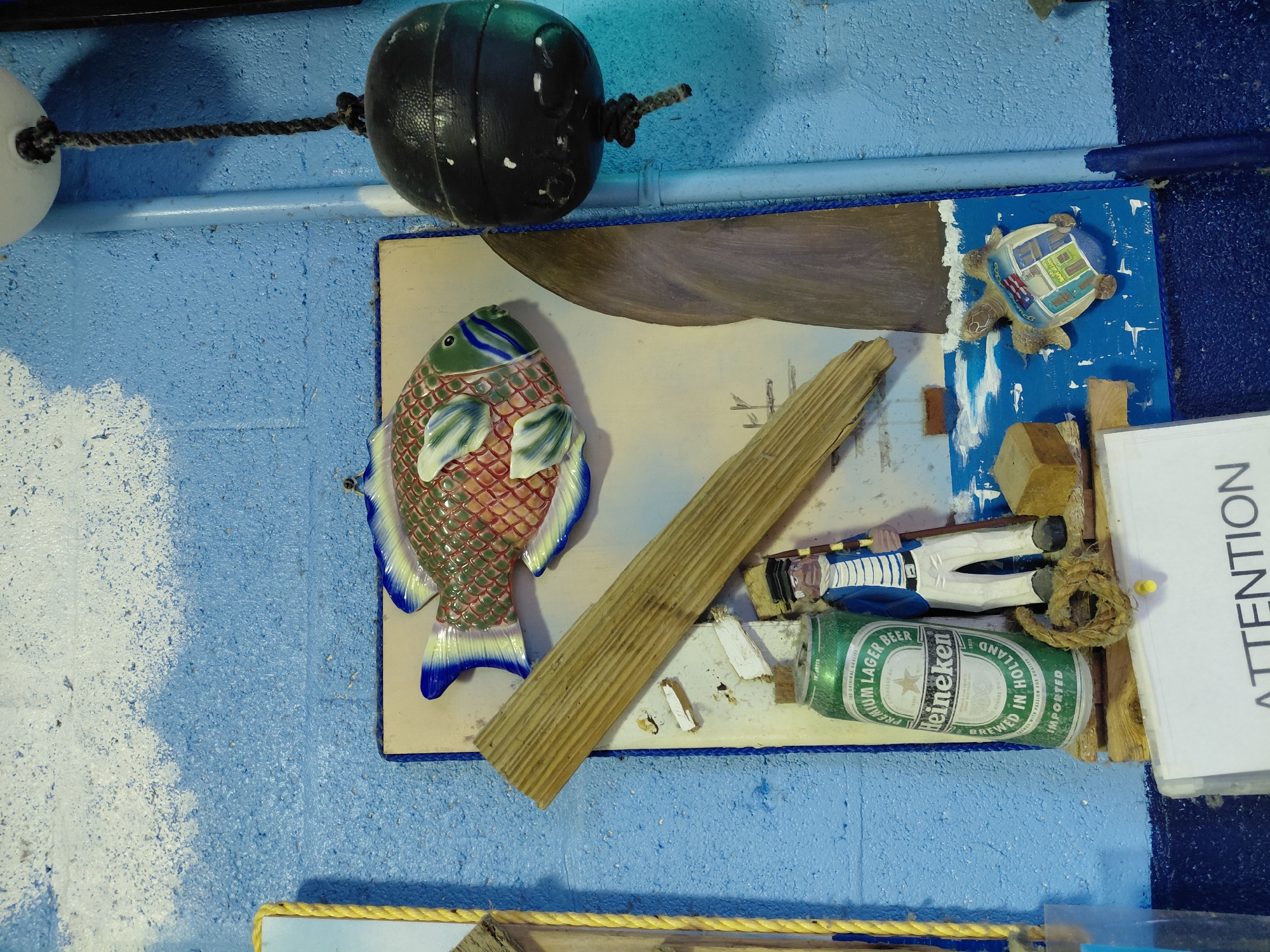 The image shows a nautical-themed display affixed to a textured blue wall. A black buoy hangs from the top. The display features a ceramic fish, a wooden plank, a miniature turtle figurine, a toy sailor, and a Heineken beer can. An "ATTENTION" sign is also present. It is likely a decorative display in Cutler Bay, Florida.