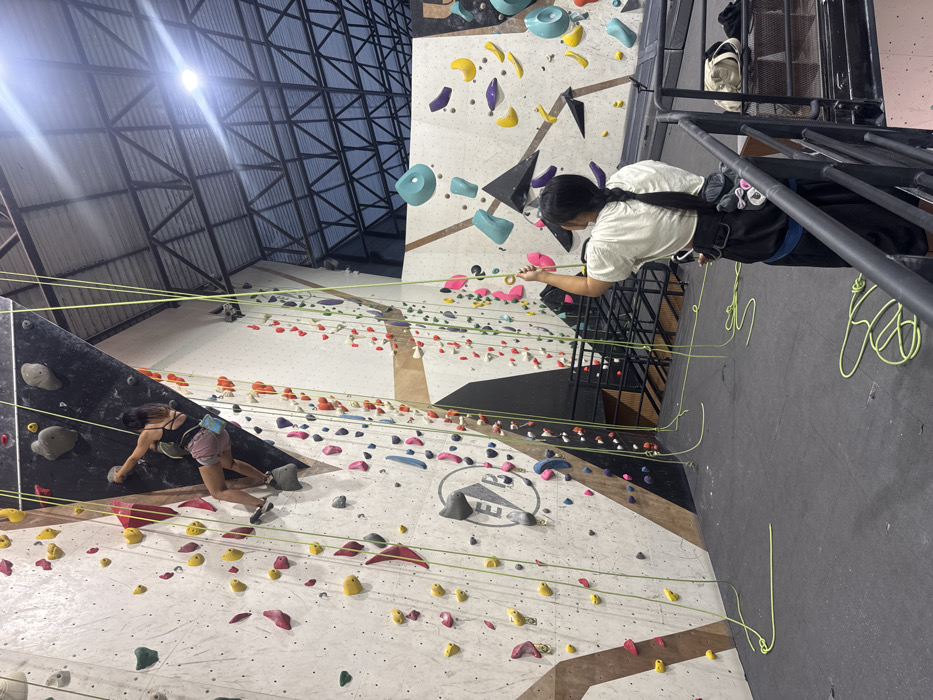 An indoor climbing gym scene at Easy Bouldering Club (EBC) in Bangkok, Thailand, features two females engaged in rock climbing activities. In the upper left quadrant, a female climber ascends a vertical wall section, approximately halfway up. She wears a black tank top, pink shorts, and climbing shoes, with a light blue chalk bag attached to her waist. Her right hand grips a dark grey hold, and her left foot is positioned on a light grey hold. Several bright yellow ropes hang vertically alongside her route. In the foreground, positioned lower and to the right, a second female stands on the padded floor, facing away from the viewer and looking towards the climbing wall. She has long dark hair styled in a braid, wears a light-colored t-shirt, dark pants, and a black climbing harness. Her right hand grasps a yellow climbing rope, indicative of belaying or rope management. Coiled yellow ropes lie on the dark grey padded floor. The climbing walls are predominantly light grey with sections of dark grey and wood-patterned accents, densely covered with numerous multi-colored holds (yellow, red, blue, green, pink, purple, grey). A prominent "EBC" logo in a grey circle is visible on a light grey wall section to the left. The high ceiling features exposed dark metal truss work and a translucent roof, with a bright overhead light visible in the upper right, emitting lens flare. Other climbing walls with distinctive large blue holds are visible in the background.