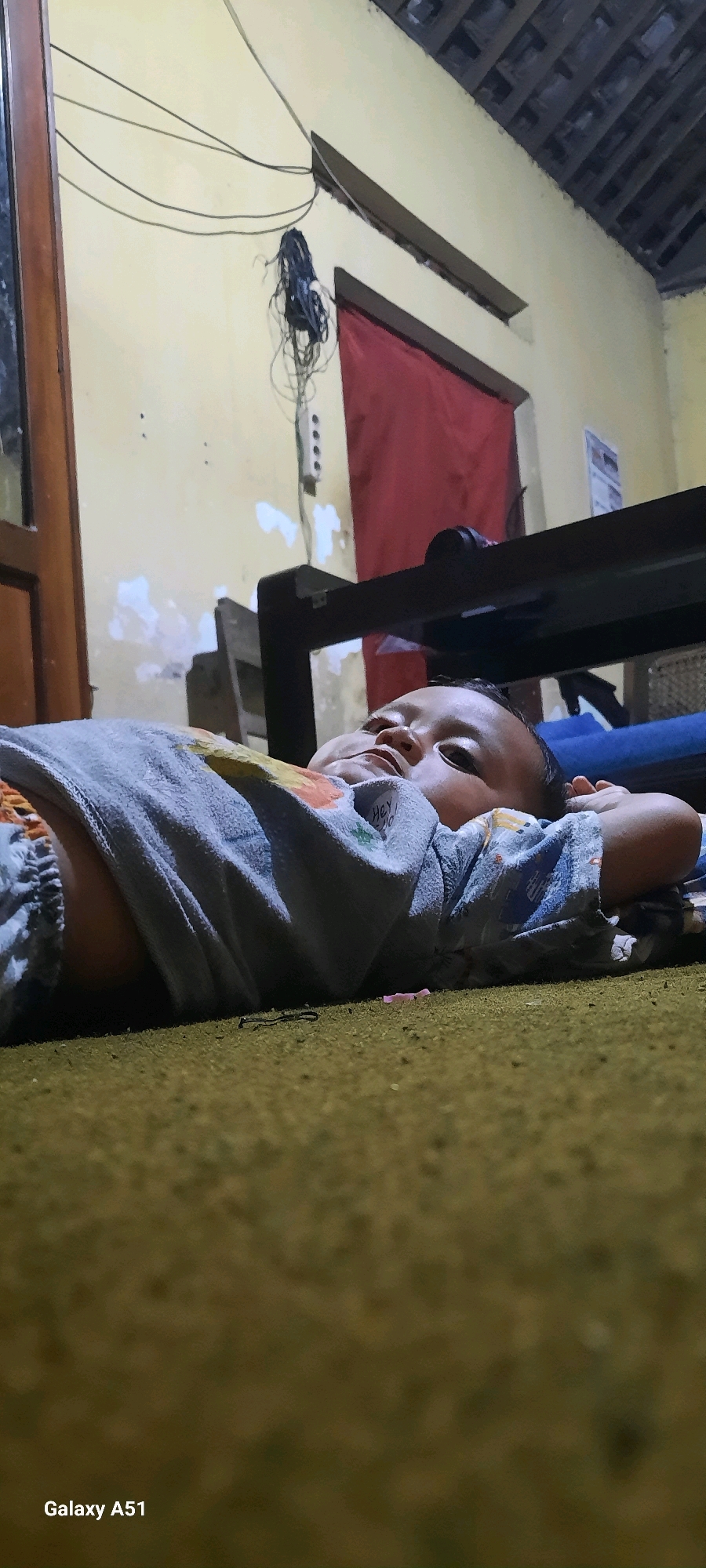 The image shows a close-up, low-angle view of a baby lying on its back on a brown carpet. The baby is wearing a grey and blue patterned pajama set. The baby's head is slightly tilted to the right, with its hands behind its head. The background is dimly lit and shows a wall painted yellow, with some peeling paint. Above the wall, a dark ceiling with exposed beams is visible. To the left, a wooden door frame can be seen. Electrical wires are hanging from the wall and bunched together. A red piece of fabric is also visible on the wall. A dark furniture piece is present behind the baby. The scene appears to be indoors, likely in a residential setting. The text "Galaxy A51" is present in the bottom left corner, indicating the device used to capture the image. The context provided suggests the location is the city of Jatitengah, Indonesia.