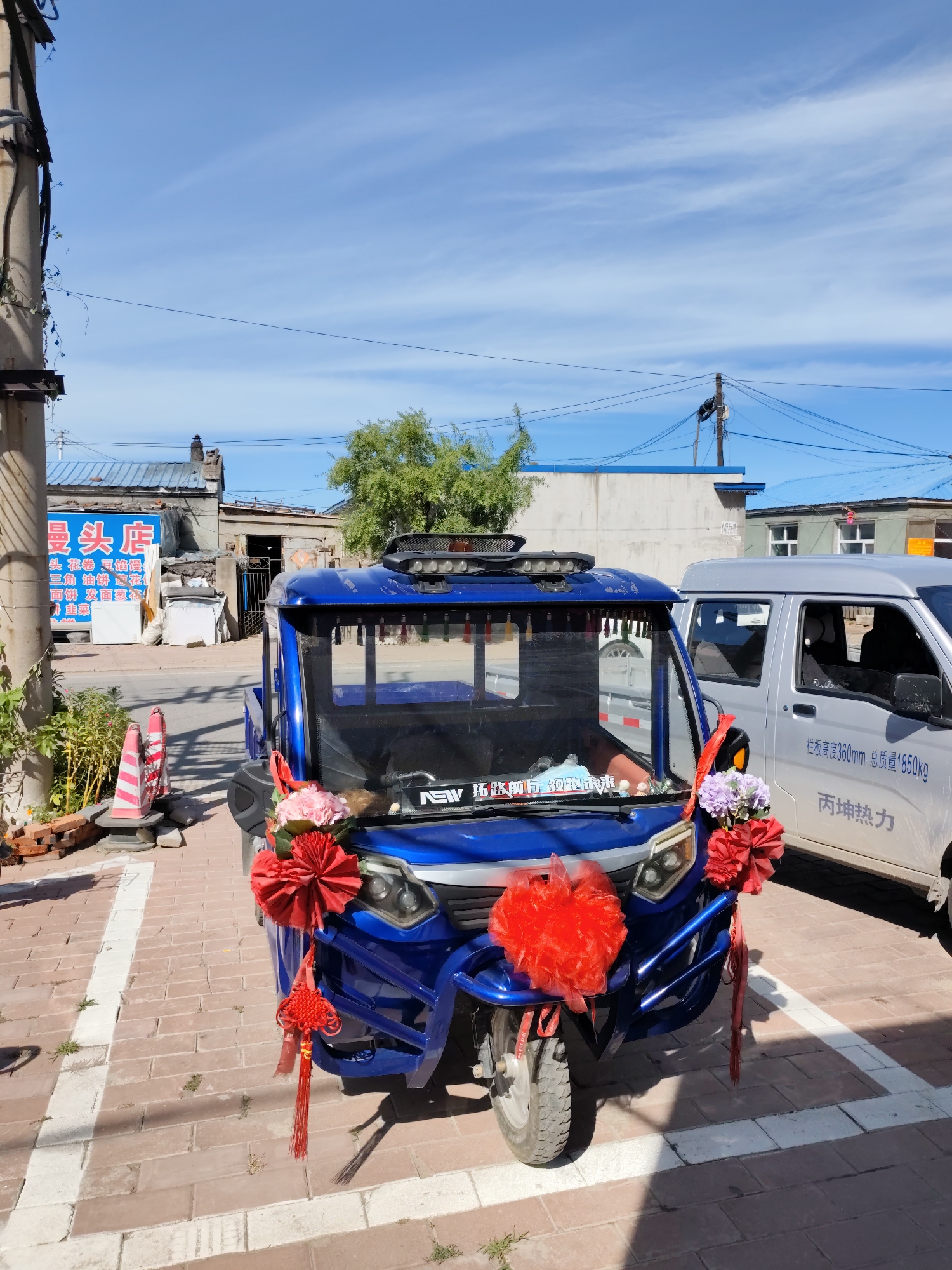 This image was taken outdoors on a sunny day in Lindian, China. In the foreground, a bright blue three-wheeled vehicle is parked on a brick-paved area marked with white lines. The vehicle is decorated with red and pink fabric bows and a red tasseled ornament. Behind the blue vehicle, a silver van is parked. In the background, there are low-rise buildings with blue roofs and a clear blue sky with wispy white clouds. A utility pole with numerous wires is visible to the left of the frame. The scene appears to be a street or a parking area in a residential or commercial district.