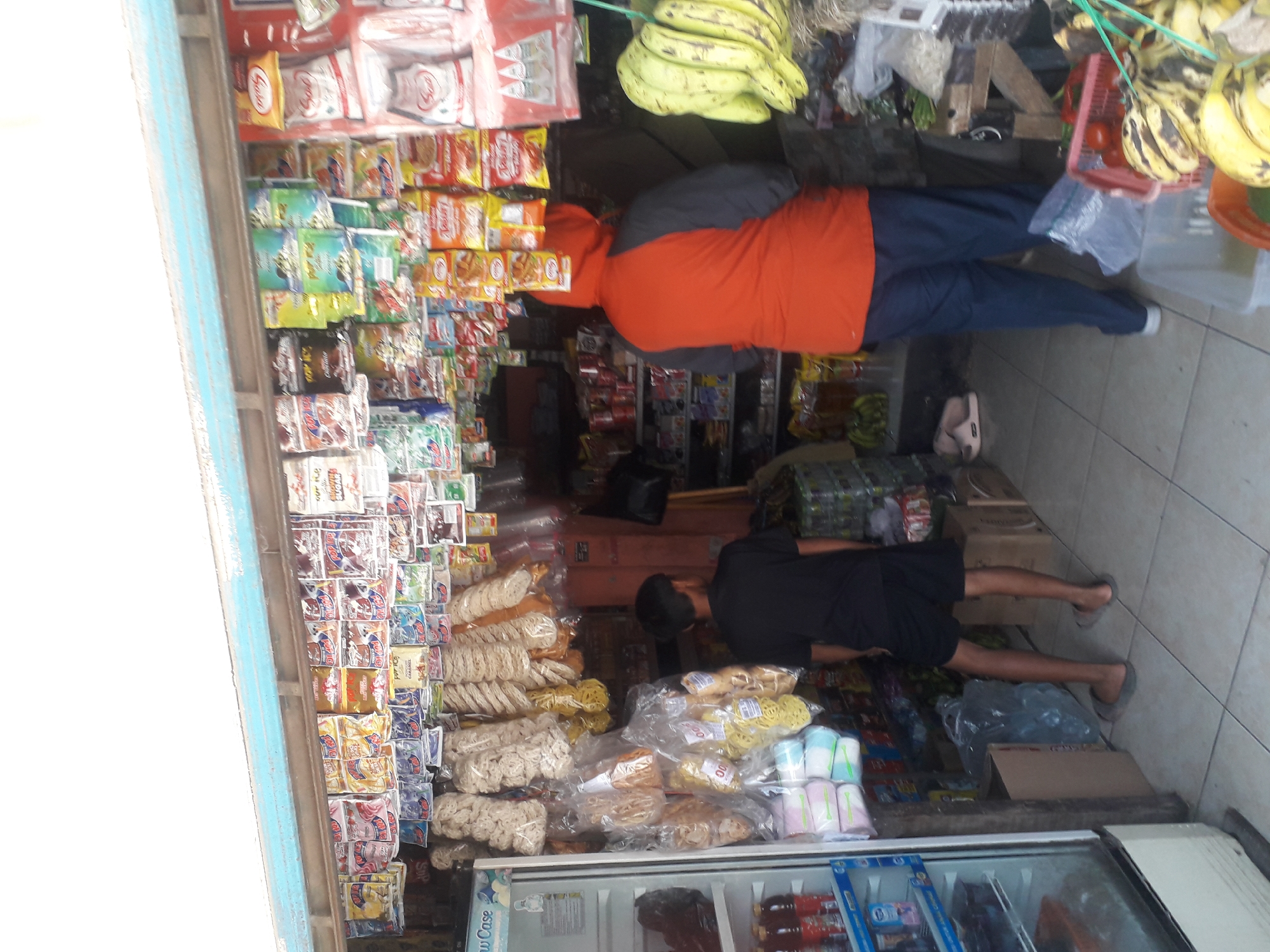 In Cankuang, Indonesia, a small convenience store is depicted. The store is packed with various goods, including packaged snacks and drinks hanging from the ceiling and displayed on shelves. A young boy in a black t-shirt and shorts stands near a refrigerator on the left. Further inside, a person wearing an orange hoodie and blue pants is visible. The floor is tiled, and the overall scene suggests a typical neighborhood store.