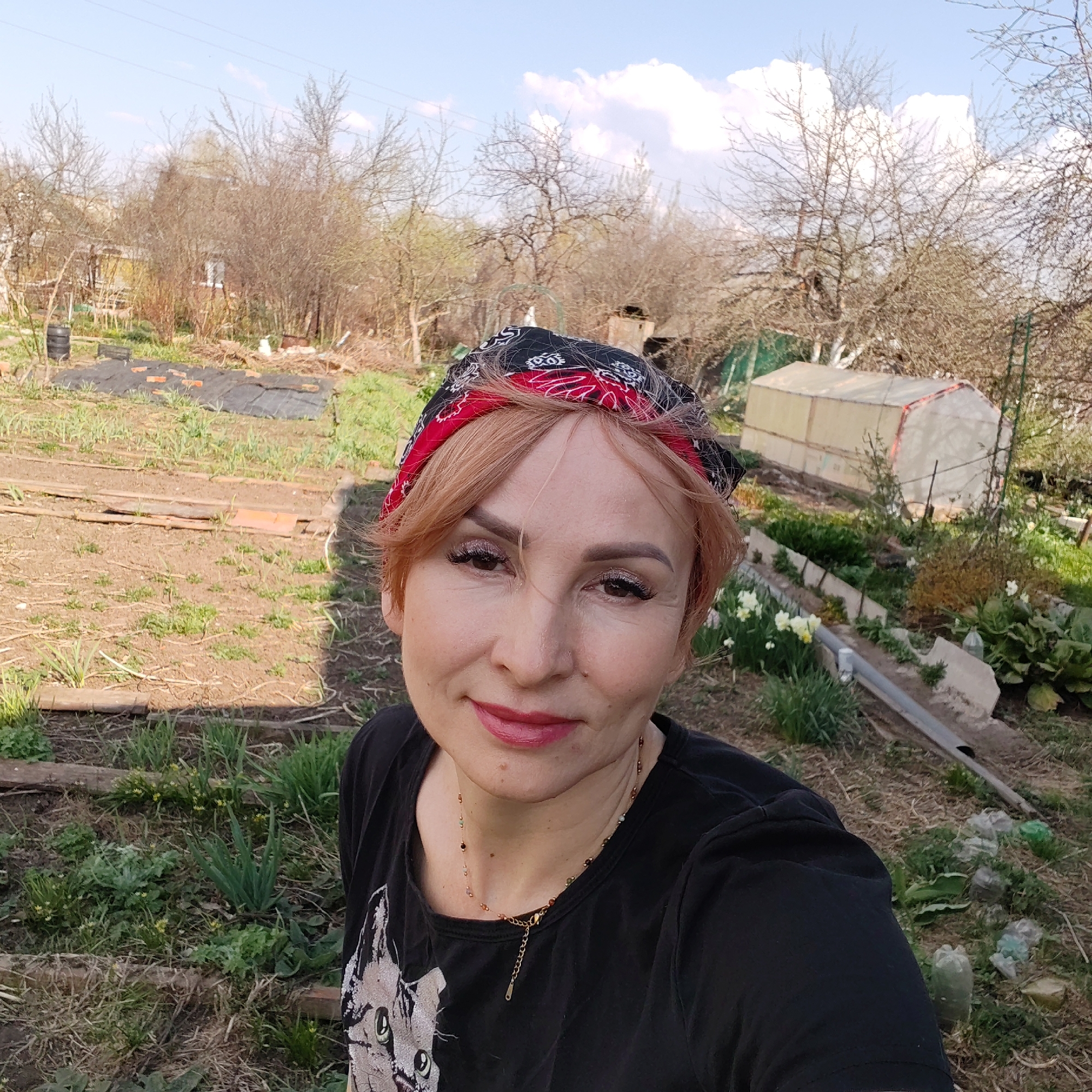 A middle-aged woman wearing a black t-shirt with a cat print and a red and black bandana stands in a garden. Her hair is short and light red. The garden has raised beds, bare trees, and a greenhouse in the background. The sky is partly cloudy. The location is Chaikovsky, Russia.