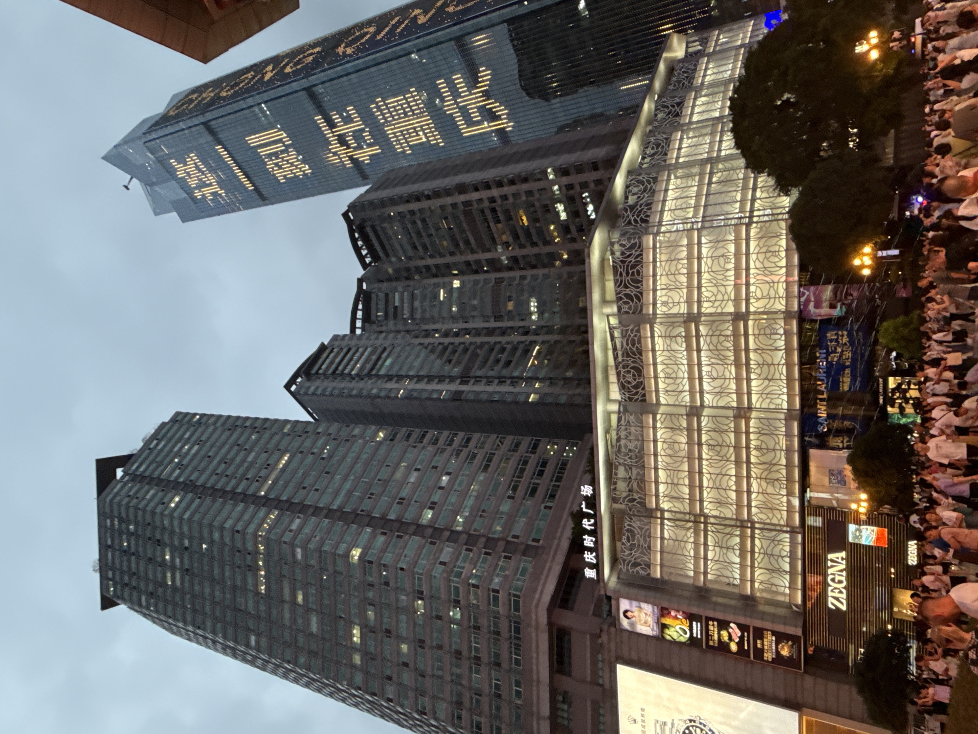This is a low-angle outdoor photograph taken at dusk or night in the Yuzhong District of Chongqing, China. The scene is dominated by several tall, modern skyscrapers. The building on the far right has large Chinese characters illuminated in yellow on its facade. In the foreground, a crowd of people is gathered on a street or plaza. Below the skyscrapers, there are brightly lit storefronts, including one with the "ZEGNA" logo and another with "SAINT LAURENT." The sky is overcast and gray, providing a diffused light that highlights the illuminated signs and building lights. The overall impression is of a busy, modern urban environment at night.