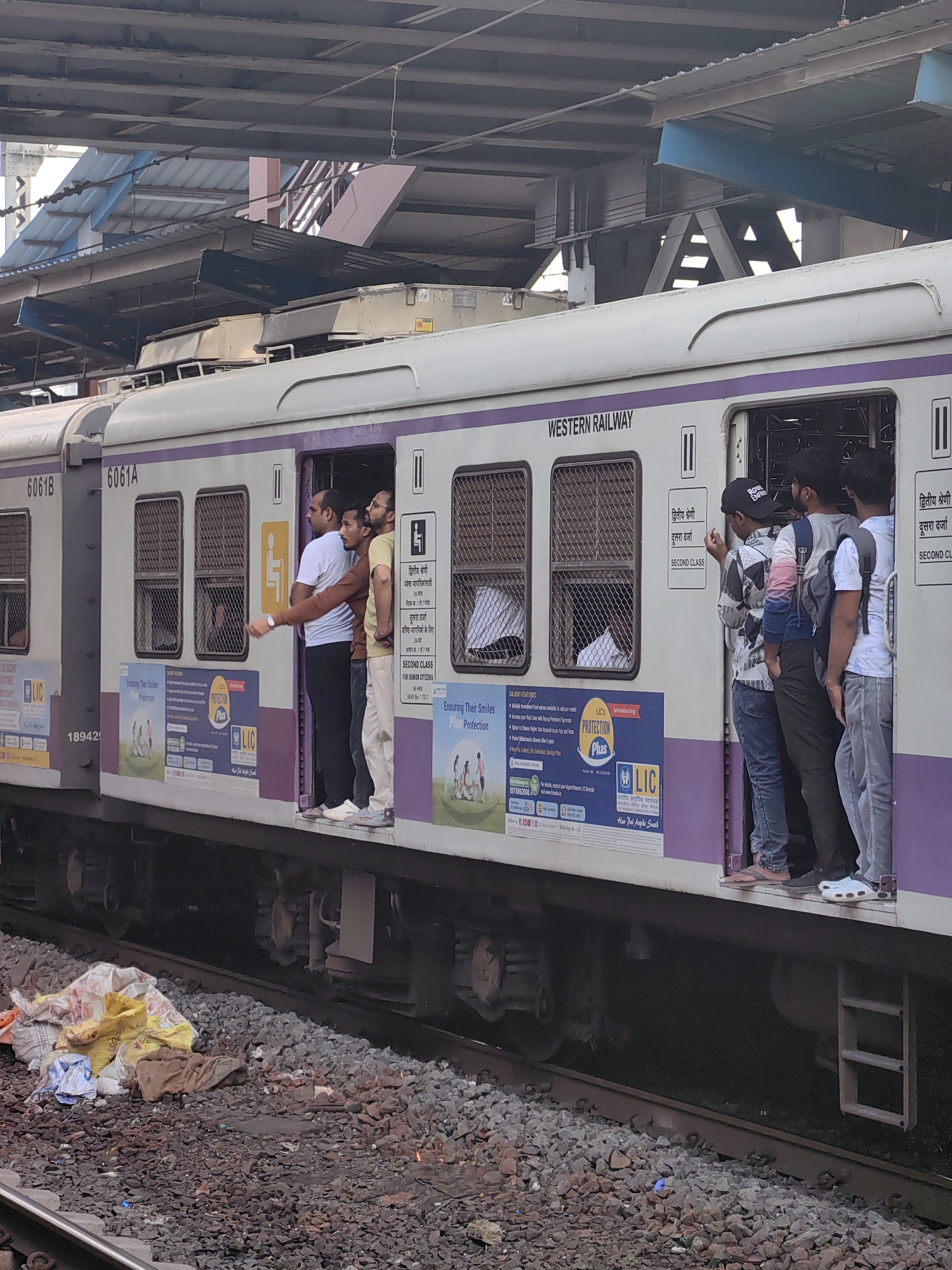 The image depicts a crowded suburban train car in Mumbai, India, with several passengers hanging out of the open doorway. The train is white with a purple stripe, and "WESTERN RAILWAY" is visible on the side. Advertisements for LIC are plastered on the train's exterior. The train is stopped at a station, as indicated by the overhead platform structure and the tracks. Passengers are standing on the train's platform and some are leaning out the door, suggesting a crowded commute. The ground around the tracks is covered in ballast and some scattered debris, including plastic bags and fabric. The overall scene conveys the characteristic image of a busy Mumbai local train during peak hours.