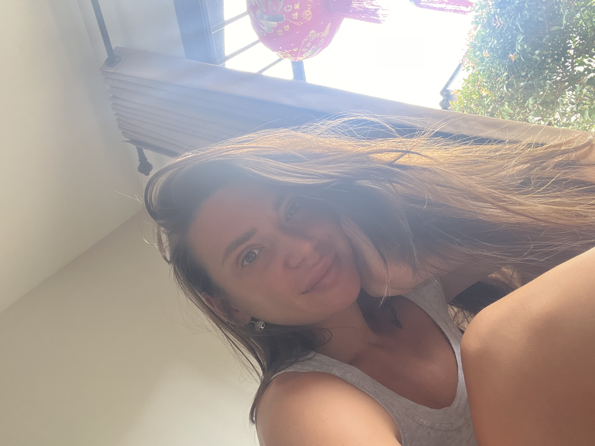 A low-angle, close-up shot depicts a woman with long brown hair, fair skin, and blue eyes. She is wearing a light gray tank top. The background is bright, suggesting daytime. A window with closed blinds is visible in the upper portion of the frame, along with a red, spherical object, possibly a lantern. Lush greenery can be seen outside the window. The location is Unknown, Unknown.