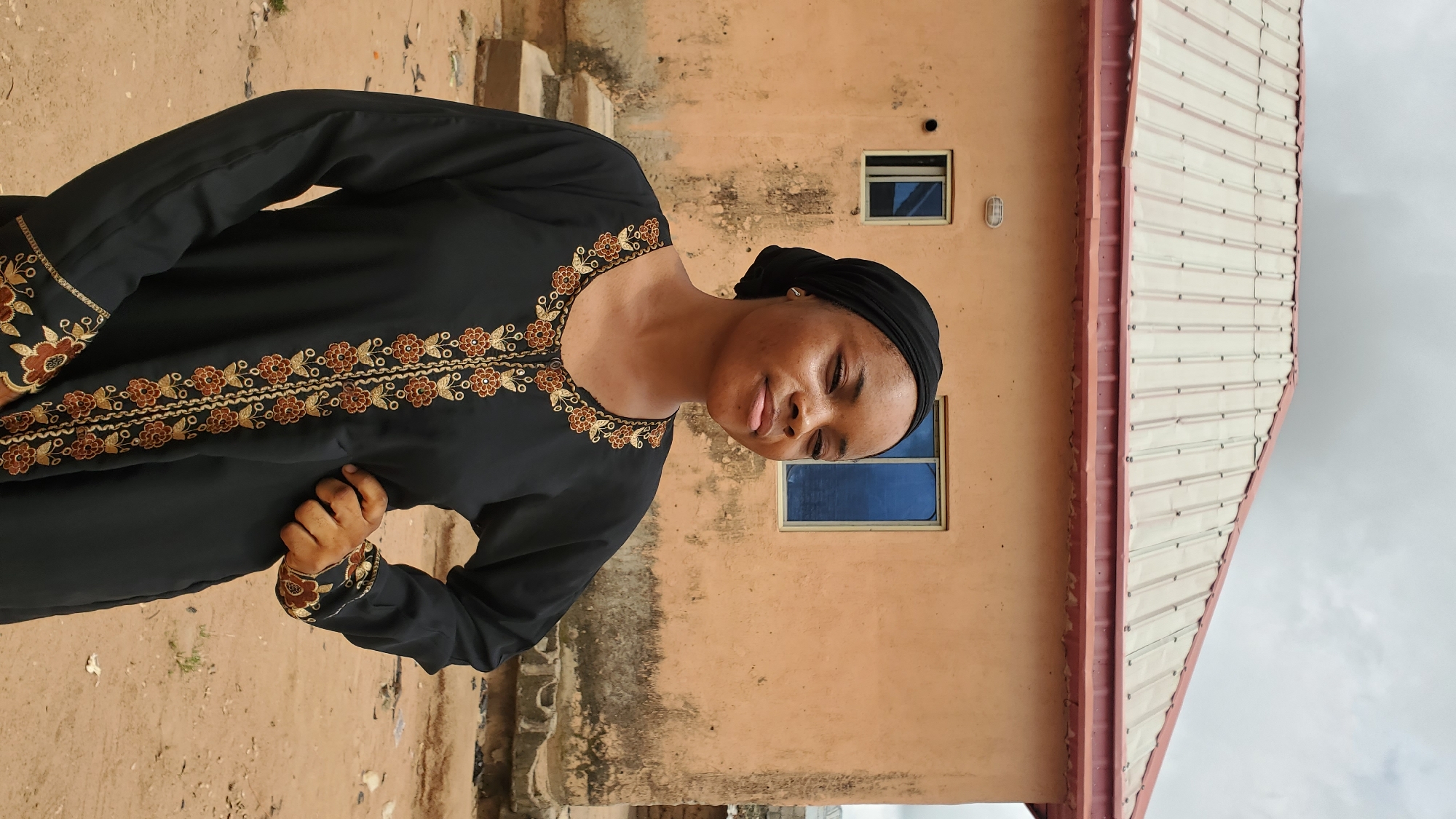A young Black woman is standing in front of a peach-colored building with a corrugated metal roof. She is wearing a black hijab and a black dress with ornate gold and brown embroidery along the neckline and cuffs. Her right hand is on her hip. The building has two small, square windows with blue glass. The ground is made of reddish-brown dirt with some scattered debris. The sky is overcast. The location is the city of Jalingo, Nigeria.
