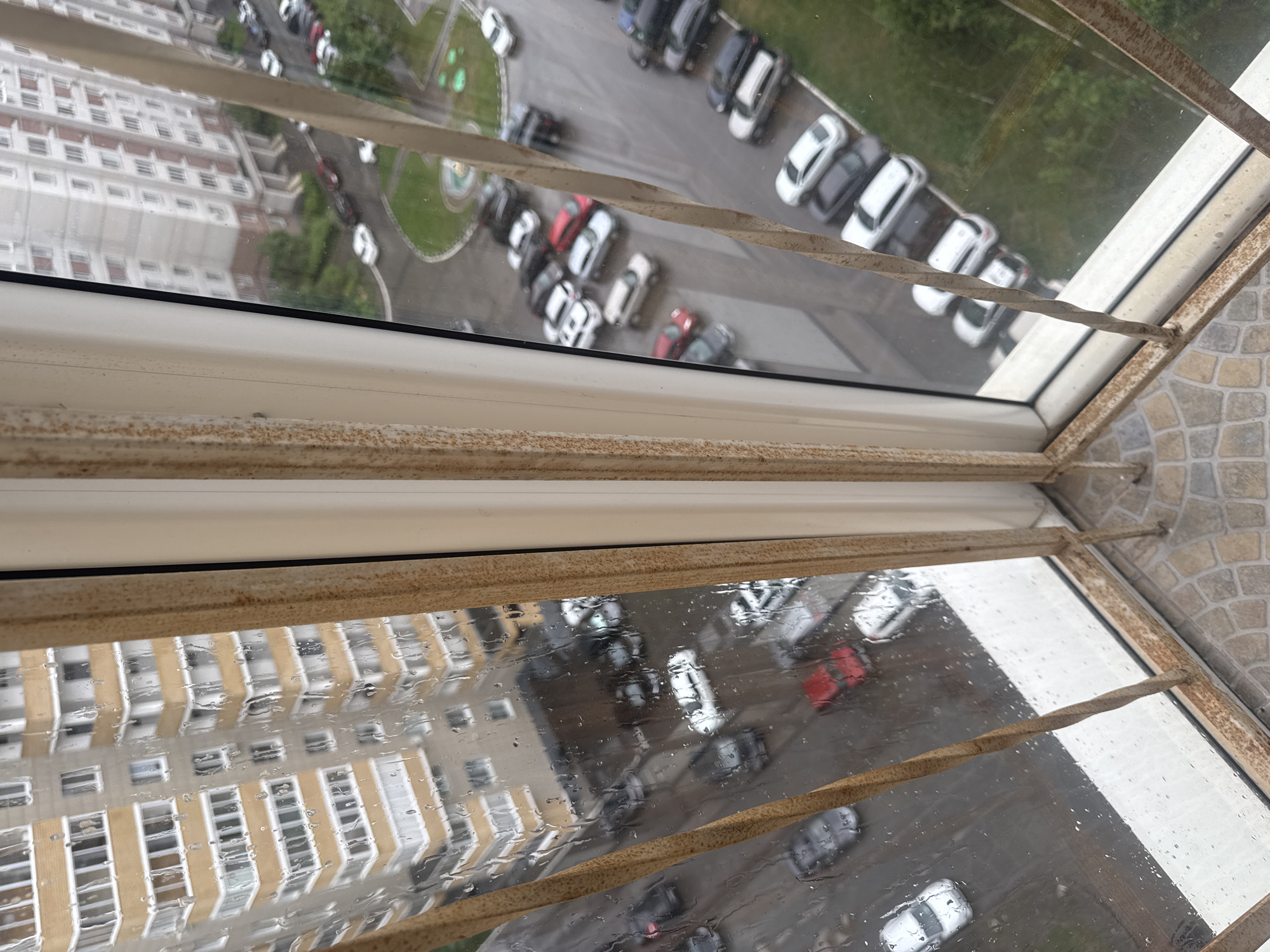 The photograph shows an elevated view of an urban environment seen through a rain-streaked window. The view includes a multi-story residential building with a light facade, a parking lot with numerous parked cars, and some greenery. The foreground features the window frame and security bars, which appear to be weathered and possibly rusted. The weather is overcast and wet.