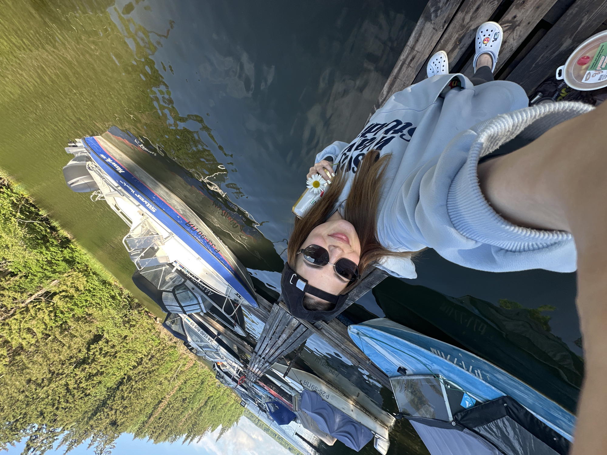 A young Caucasian woman wearing a black baseball cap, sunglasses, and a light blue hoodie is taking a selfie while standing on a wooden dock. She is holding a can and a white daisy. She is wearing white Crocs. Several boats are docked on the lake, with a treeline in the background. The image was taken in Zhebrei, Russia.