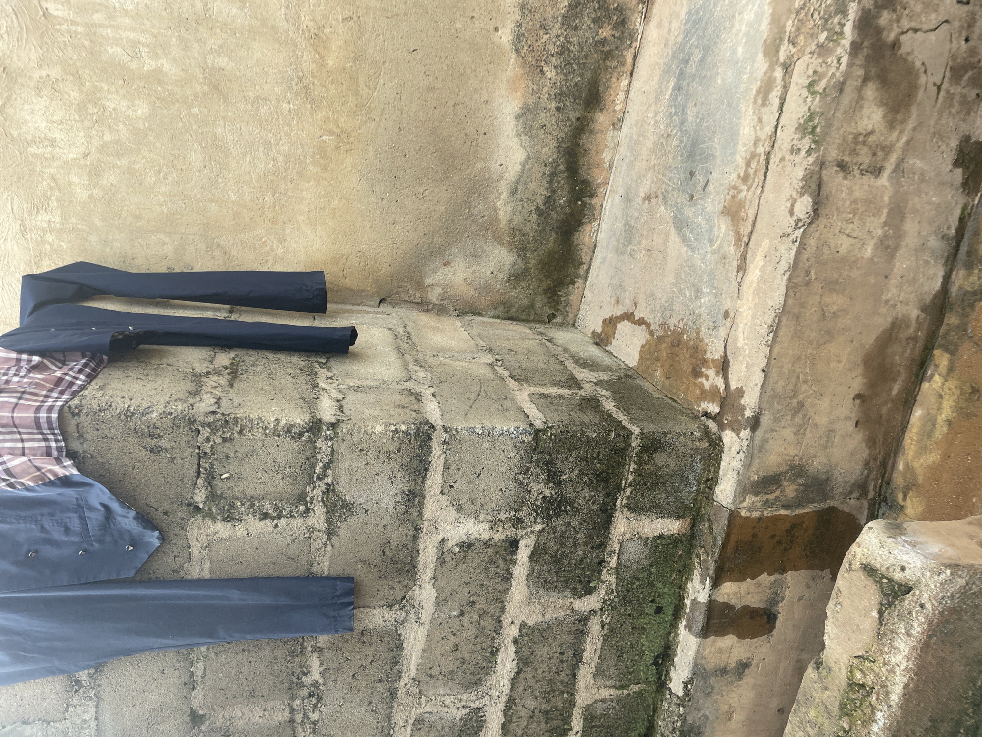 The image shows a corner of a brick wall in Lokoja, Nigeria, with some clothing hanging on it. The wall is constructed of cinder blocks and shows signs of age, with moss and water stains. A patterned shirt and dark blue pants are draped over the corner of the wall. Below the wall, a concrete ledge is visible, stained with water and dirt. The ground appears to be a channel or trough, also filled with murky water. The overall impression is of an outdoor area, likely a yard or alleyway, where laundry is being dried. The texture of the concrete and brick, along with the water stains and moss, suggests a humid environment.