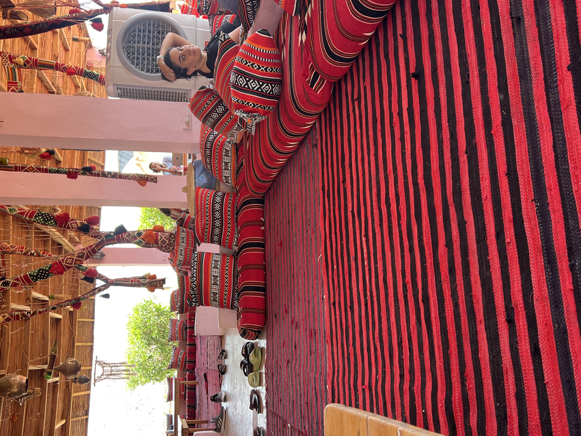 The image depicts a woman resting on floor cushions adorned with red and black striped patterns and intricate geometric designs. She is leaning against a larger floor cushion, with her hand resting on her head, looking directly at the camera. The scene appears to be an outdoor seating area, likely a cafe or lounge, with a thatched roof overhead. Various decorative elements, including fabric-draped poles with tassels and hanging metal lanterns, are visible. In the background, a large white cooler unit is present. The floor is covered with a thick, red and black striped rug. Several pairs of sandals are scattered on the floor to the left. The overall ambiance suggests a relaxed, traditional setting in Nueiba, Egypt, possibly a beachside establishment.