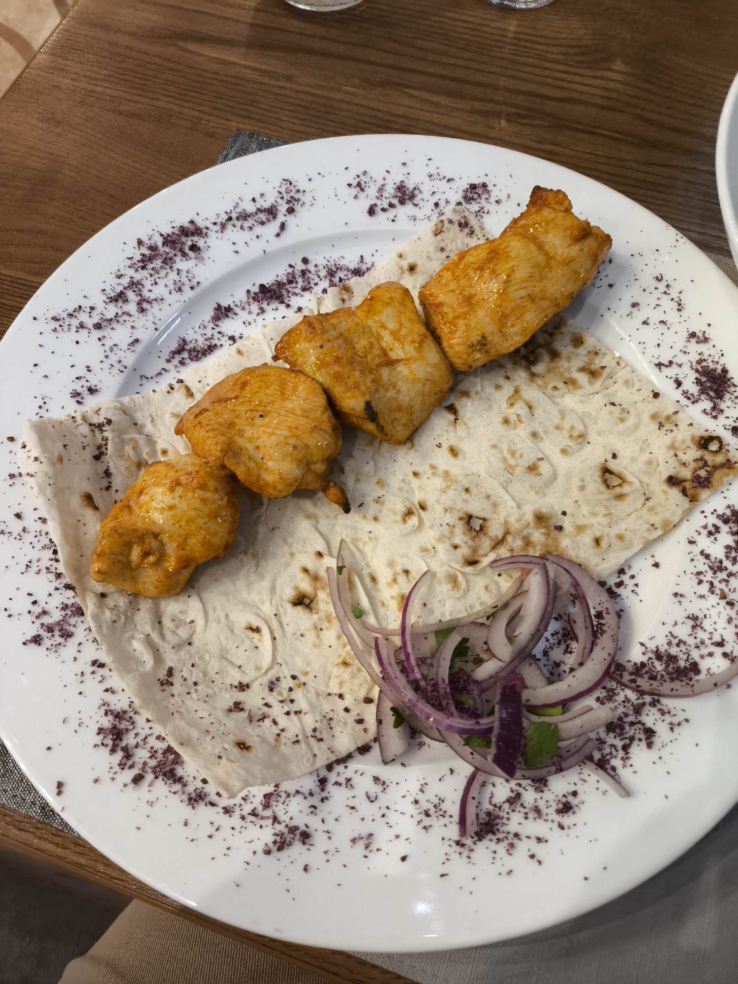 A white, oval-shaped plate filled with a meal is captured in a high-angle, close-up shot. The main subject is a serving of chicken shish kebab, featuring four pieces of golden-brown, marinated chicken. These chicken pieces are laid on a piece of light-colored flatbread, resembling lavash, which displays some darker, slightly charred marks.
To the lower right of the chicken and flatbread, a garnish of thinly sliced red onions and a scattering of chopped green herbs, likely parsley, is present. A dark reddish-purple spice, consistent with sumac, is sprinkled around the rim of the plate and over parts of the flatbread, enhancing the visual appeal.
The plate is set on a dark wooden table. A grey fabric, possibly a placemat or napkin, is partially visible beneath the plate on the right side. In the blurred background, hints of a clear glass and another white dish can be seen. The image depicts a prepared meal, possibly served in a restaurant or home dining setting in Khimki, Russia. No people or text are visible.