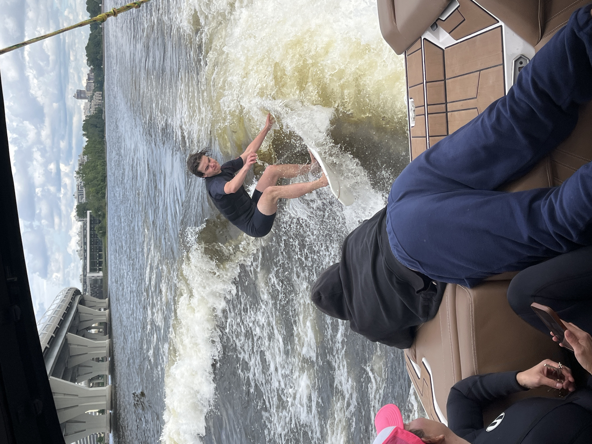 In Saint Petersburg, Russia, a person is wakeboarding on a body of water, likely the Neva River, given the urban landscape and bridge visible in the background. The person is wearing a black wetsuit and is mid-action, carving through the water and creating a large wake. The boat from which the photo is taken is to the right, with passengers visible, one of whom is wearing blue pants and another is holding a phone. The sky is cloudy, suggesting it's daytime, possibly overcast. The overall mood seems to be one of recreation and enjoyment.