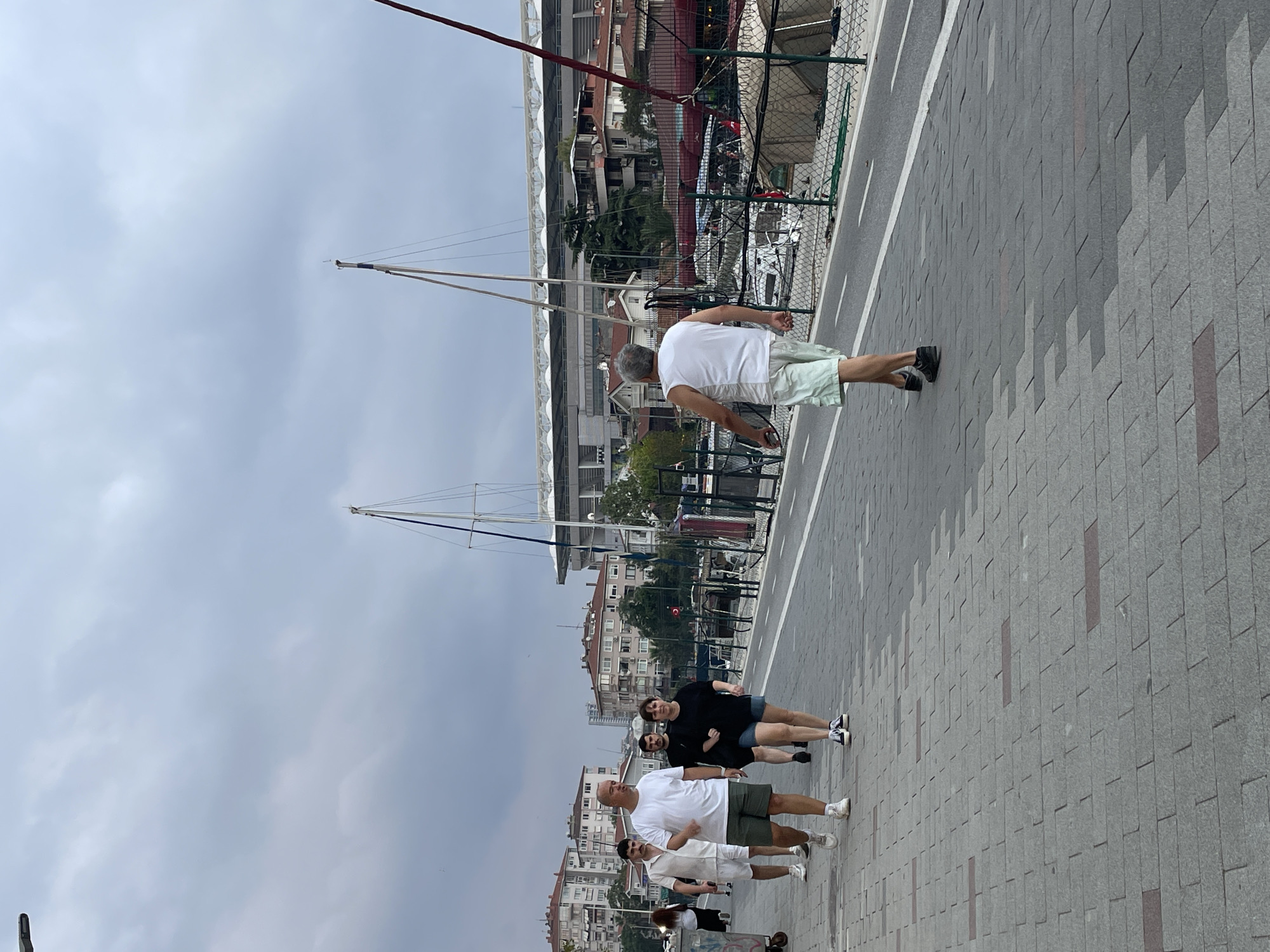 This is a full-frame, eye-level, outdoor, vertical, medium-distance shot of a cloudy day on a pedestrian walkway. The walkway is paved with interlocking grey and brown rectangular stones. The scene appears to be along a waterfront in Kadıköy, Turkey, with buildings, a stadium, and boats visible in the background. Several people are walking on the path. In the foreground, a man in a white tank top and light green shorts walks away from the camera. Further ahead and to the left, a group of four people walk towards the camera: a man in a white t-shirt and green shorts, a man in a black t-shirt and dark shorts, and a woman in a black t-shirt and denim shorts. Another man in a white short-sleeved shirt and white shorts is walking further left and slightly ahead of this group. A white garbage bin is visible on the far left. In the background, there are several multi-story buildings with red-tiled roofs. A large stadium with a white canopy is prominent in the middle-ground, and several boats and masts are visible along the water's edge. A fence with a green net runs along the right side of the walkway. The sky is overcast with dark grey clouds.