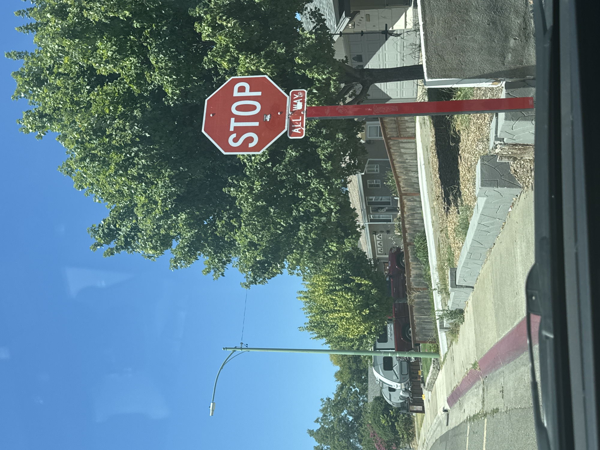 The image captures a street scene in Citrus Heights, California. A red, octagonal STOP sign with a white border and white lettering is prominently displayed on a red metal post. Below the STOP sign, a smaller rectangular sign with a white border and red lettering reads "ALL WAY." The STOP sign is positioned to the right of a light green street lamp with a curved arm.
In the background, a large, leafy green tree dominates the right side of the frame. To the left of the tree and behind the STOP sign, a residential house with a light-colored facade and a garage door is partially visible. In the mid-ground, a sidewalk runs along the street, bordered by a low retaining wall constructed from grey, irregularly shaped concrete blocks. Beyond the sidewalk, there are parked vehicles, including two recreational vehicles (RVs) and a red pickup truck. Further in the distance, more houses and trees are visible under a clear blue sky. The foreground shows the dashboard of a vehicle, suggesting the image was taken from inside a car. The road in the foreground has a red-painted curb.