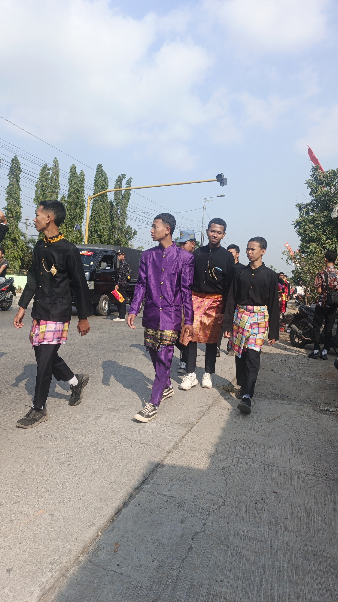 In Sumengko, Indonesia, a group of young men in traditional attire are walking down a paved street under a bright, partly cloudy sky. The individuals, all appearing to be teenagers or young adults, are dressed in long-sleeved shirts and pants, some adorned with sarongs or sashes. One young man in the center wears a vibrant purple outfit, while others are in black with colorful patterned sashes or sarongs. They appear to be participating in a procession or parade, moving from left to right across the frame.
In the background, a black truck is visible, along with trees lining the street and power lines overhead. There are also motorcycles parked on the right side of the street. The road surface is a light gray concrete with visible cracks and lines. The lighting suggests it is daytime, with shadows cast by the individuals and trees onto the street, indicating the sun is positioned to the left. The overall scene is one of an outdoor community event or celebration.