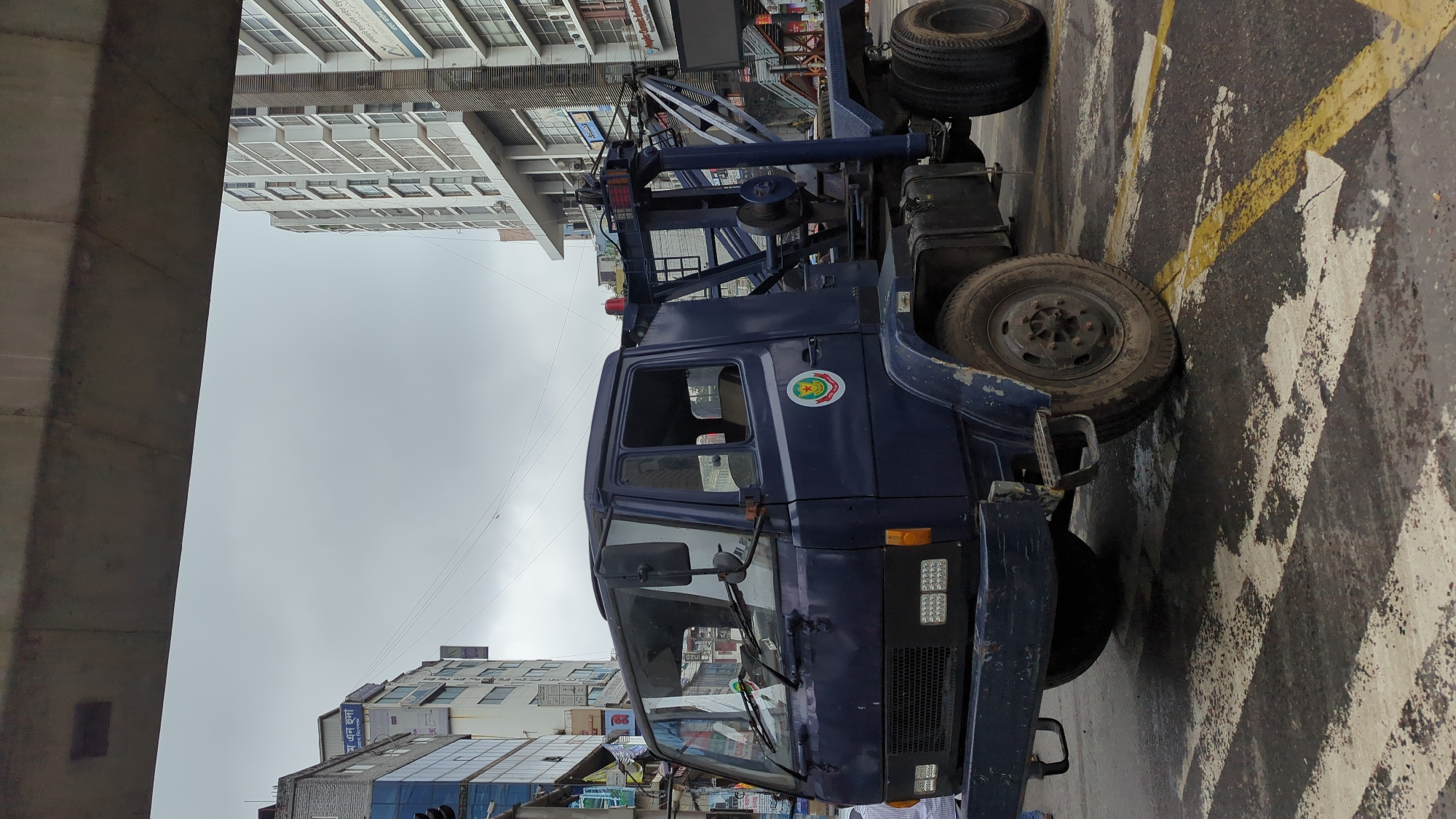 A dark blue tow truck, likely belonging to a government agency given the emblem on the door, is parked on a street in Dhaka, Bangladesh. The truck is positioned on a road with yellow and white markings, possibly a crosswalk or lane divider, which appears wet and somewhat worn. In the background, modern multi-story buildings rise against an overcast sky. The truck's boom is raised, indicating it is ready for or has recently completed a task. The overall scene suggests an urban environment with ongoing city activity.