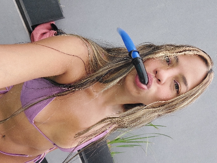 A woman with blonde-streaked hair and a blue snorkel is positioned at an angle in the frame. She wears a purple bikini and has an olive skin tone. A glimpse of what appears to be a plant is at the bottom-right. The background is a mix of gray and black, including a patch of pink cloth.