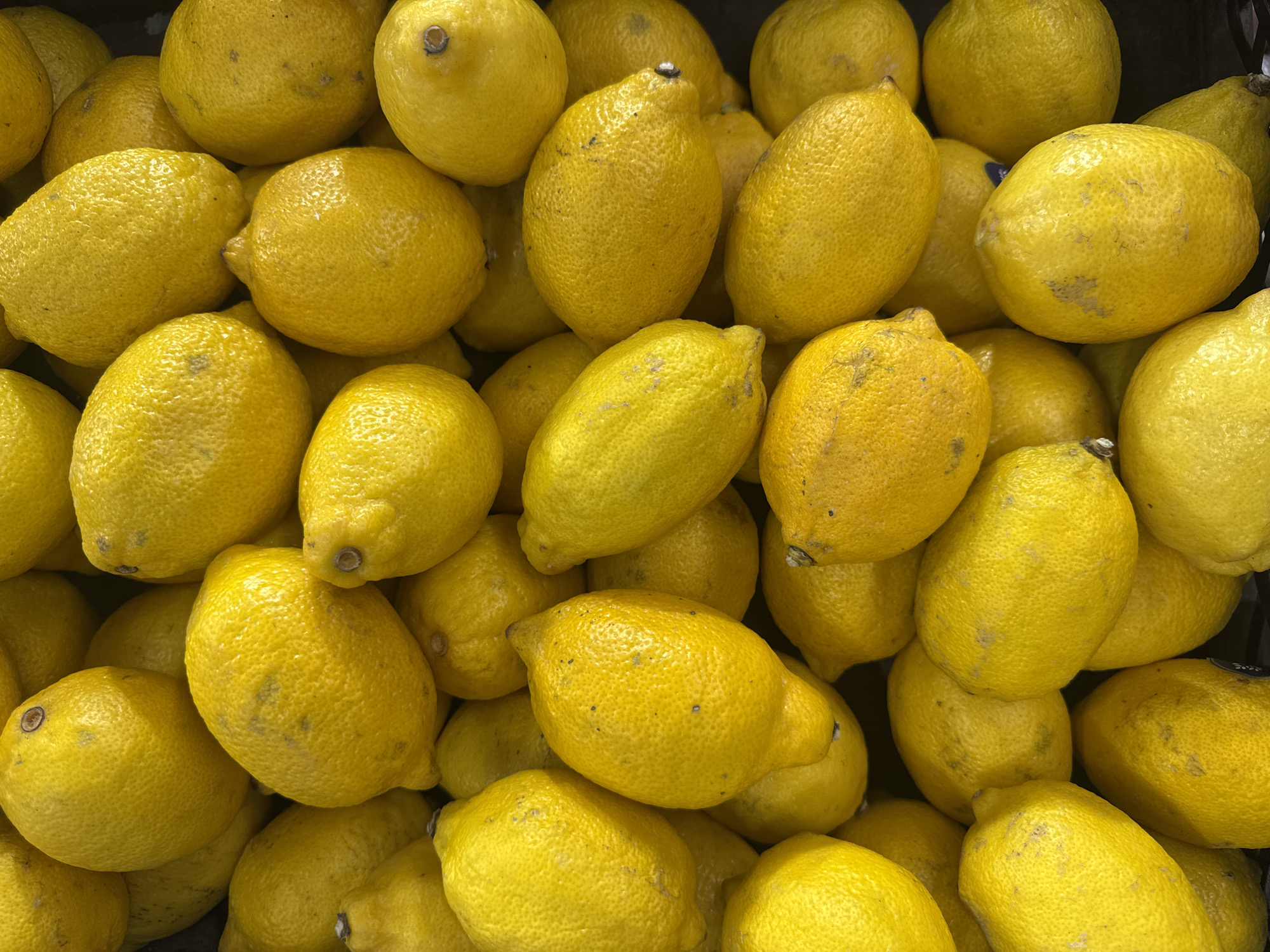 A close-up, overhead view of a pile of ripe, yellow lemons. The lemons are densely packed, creating a textured surface of bumpy, yellow skin. Some lemons show small brown spots or blemishes, indicating natural variation. The lighting is bright and even, highlighting the vibrant yellow hue of the fruit. The scene is devoid of any human presence or discernible background elements, suggesting the lemons are either in a market stall or a collection for sale. There is no indication of the specific location within Yekaterinburg, Russia, as the image focuses solely on the fruit.