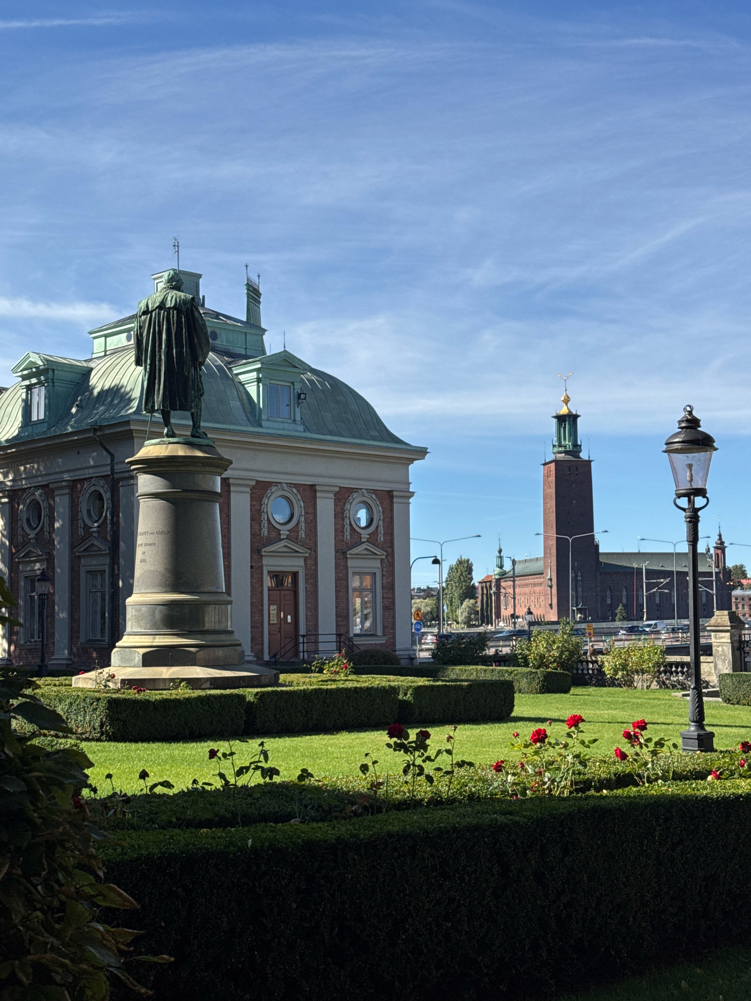The image depicts a scenic view of the Stockholm City Hall (Stadshuset) in Stockholm, Sweden. In the foreground, a statue stands on a pedestal, surrounded by manicured green hedges and a vibrant patch of red roses. To the left, a building with a green copper roof and classical architectural features is visible. In the background, the iconic tower of the Stockholm City Hall dominates the skyline, characterized by its brick facade and a green spire topped with a golden crown. The sky is clear blue with sparse white clouds, indicating a sunny day. A street with vehicles and streetlights is situated between the foreground and the City Hall. The overall scene conveys a sense of grandeur and historical significance.