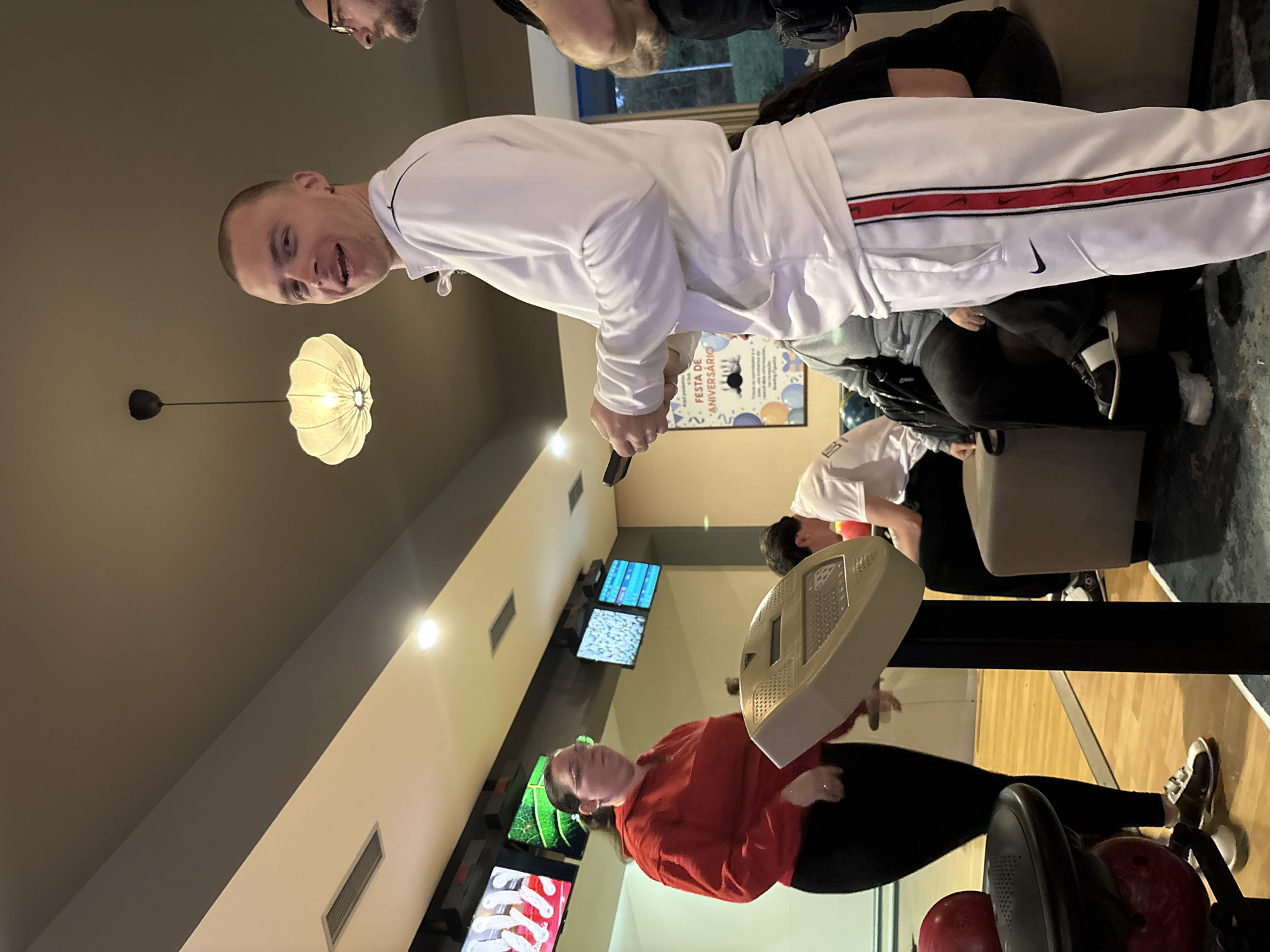 The image shows an indoor bowling alley. There are several people present, including one wearing a white tracksuit with a red stripe and a Nike logo. The bowling alley has typical features like scoring screens, ball racks, and seating. A woman in a red shirt is near the bowling lane. A light fixture hangs from the ceiling. A poster is visible on the wall. The specific city and other precise location details are not discernible from the image.