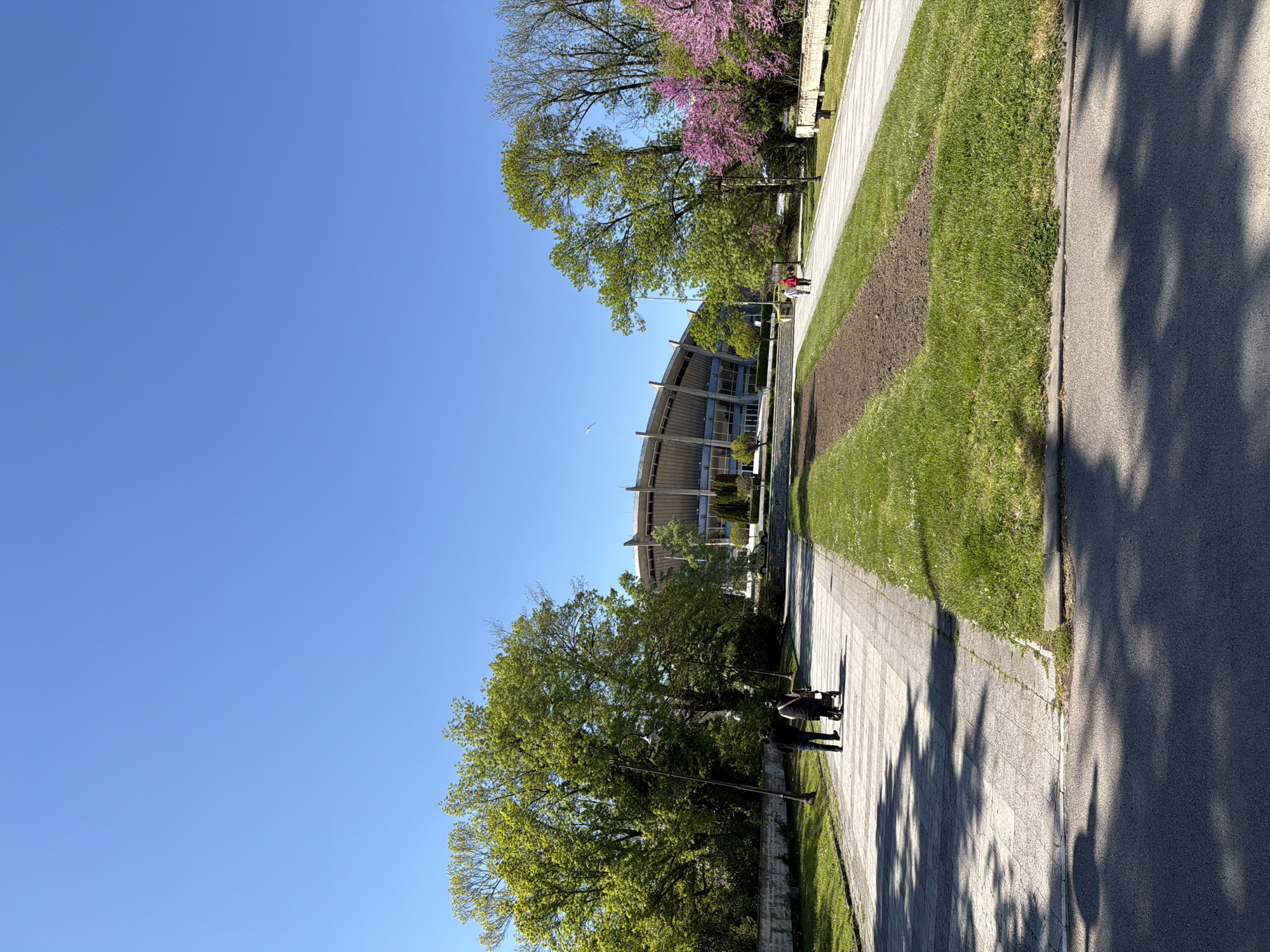 The image shows a street scene in Varna, Bulgaria on a sunny day. The sky is clear and blue. A modern building with curved elements is visible in the background. A sidewalk runs along a green lawn and a street. People are walking along the sidewalk; at least one person is wearing a red jacket. Trees are lining the path; one tree has purple flowers. Shadows from the trees are cast on the sidewalk and street.