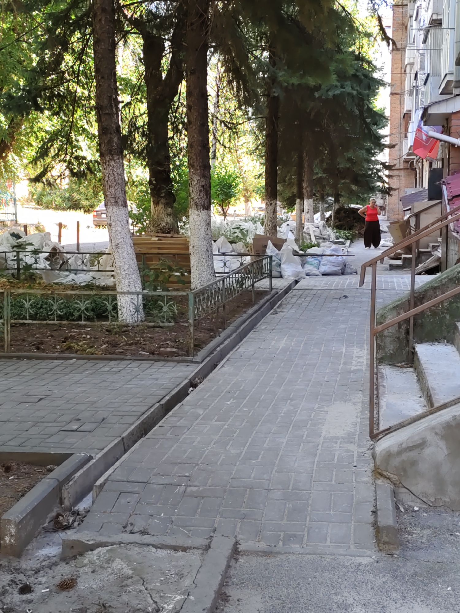 A low-angle medium shot captures an outdoor scene on a sunny day, likely in Rostov-on-Don, Russia. A woman in a red tank top and dark, wide-legged pants stands near a brick building with visible windows and a red roof. She is looking down and holding a phone to her ear. In front of the building, a ramp made of grey paving stones slopes downwards towards the viewer. To the left of the ramp is a raised garden bed with a decorative metal fence. Several trees with white-painted trunks line the path, casting shadows. Piles of white construction bags are visible behind the fence. To the right of the ramp, a concrete staircase with a metal handrail ascends. The ground is paved with grey interlocking stones, and debris, including pinecones and cigarette butts, is scattered on the ground in places. The scene appears to be an entrance to an apartment building, with construction or renovation work in progress.