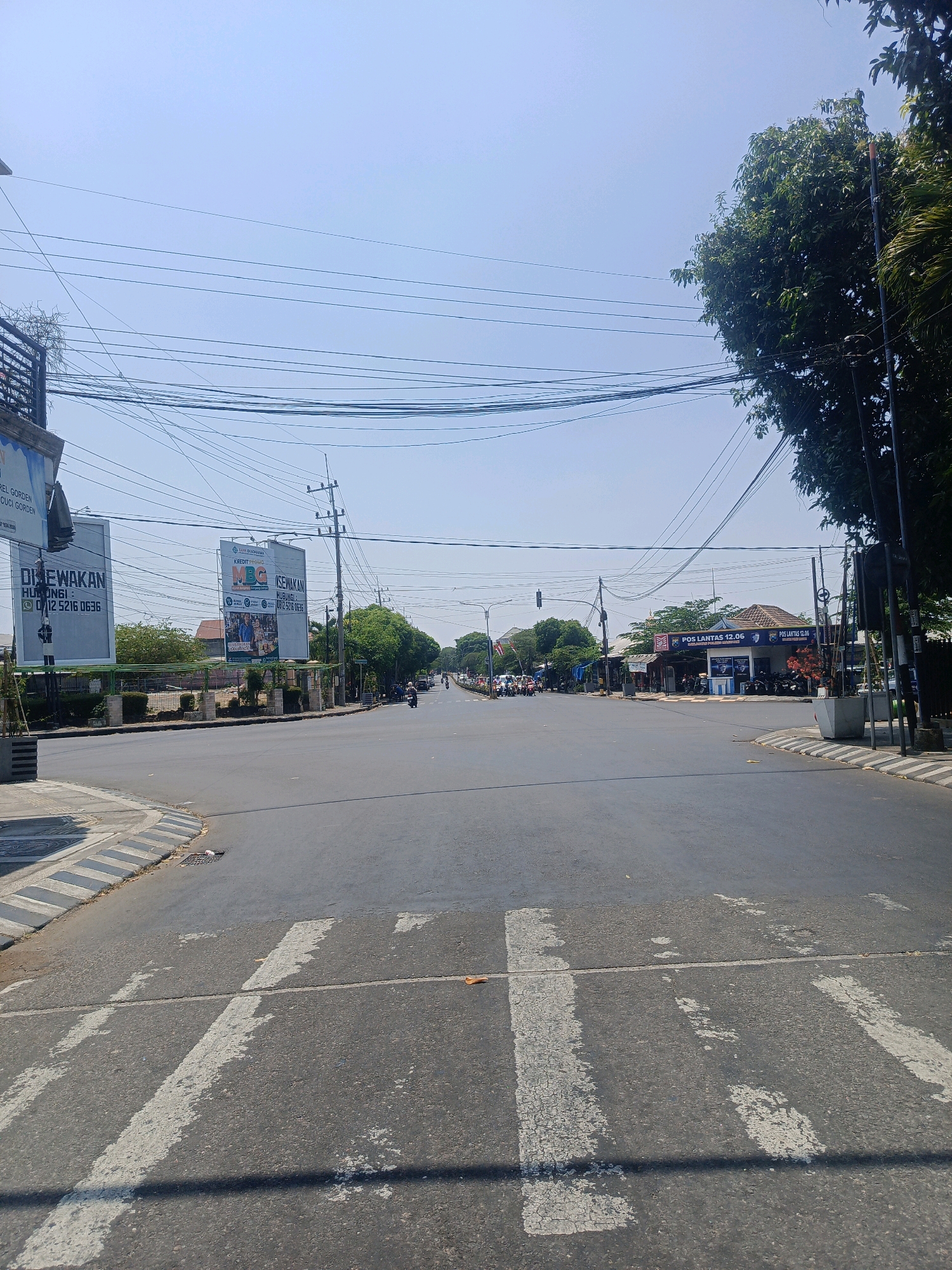 This is a wide, outdoor, eye-level shot of a road intersection in Sukowinangun, Indonesia, on a clear, sunny day. The road surface is asphalt, with clear white pedestrian crossing lines in the foreground. To the left of the road, there is a sidewalk and a small green area with some plants and a metal railing. Further back on the left, there is a large billboard. In the center of the frame, the road stretches out, with a few cars and motorcycles visible in the distance. There are power lines strung across the sky. On the right side of the road, there are buildings, including a structure with the words "POS LANTAS 12.06" on it, which appears to be a police post or traffic station. Trees with green foliage line the sides of the road in the distance. The sky is a pale blue, with no clouds visible. The overall impression is of a busy, but calm, urban intersection.