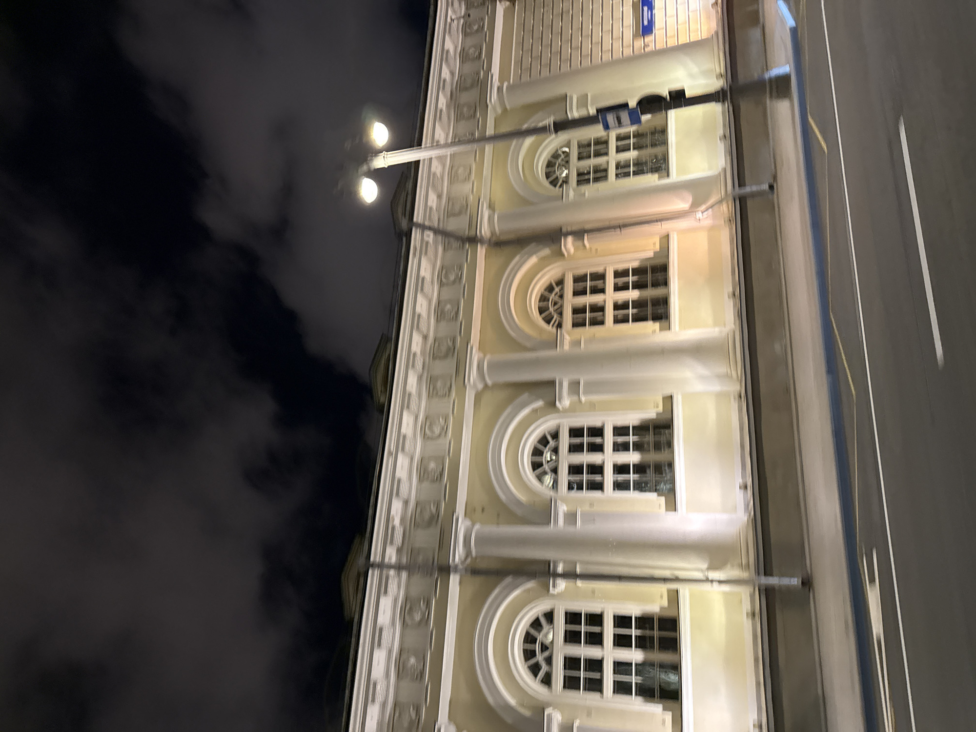 The image depicts a building exterior at night in Moscow, Russia. The building features a light yellow facade with large, arched windows, each divided into multiple panes by white mullions. The windows are illuminated from within, casting a warm glow. A prominent cornice runs along the top of the facade, adorned with decorative carvings. Several large, white columns support the cornice and are evenly spaced between the windows. A streetlamp with two illuminated globes stands on the right side of the image, casting light onto the building and surrounding area. A blue traffic sign is attached to the streetlamp post. The street in the foreground is paved and shows white lane markings. The sky above is dark with clouds. The image appears to be taken from a moving vehicle, indicated by the motion blur.