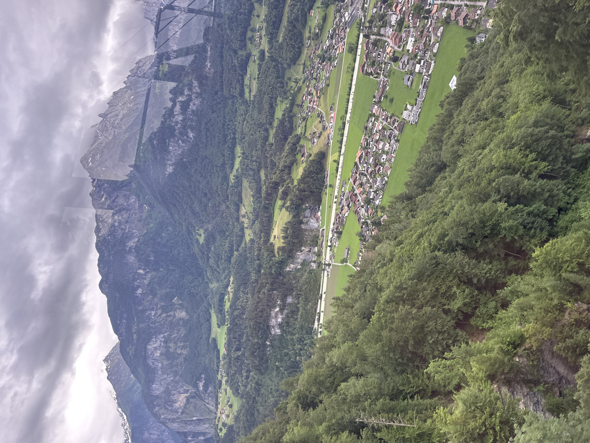 The image depicts a panoramic view of a mountainous region in or around Hüsen, Switzerland, on an overcast day. In the foreground, a steep, densely forested mountainside descends sharply. The trees are predominantly evergreen, with varied shades of green. The middle ground reveals a valley with a small village, characterized by numerous residential buildings with red-tiled roofs, interspersed with green fields and patches of forest. A road or pathway is visible, weaving through the village and fields. In the background, imposing, rugged mountains with rocky outcrops and some snow-capped peaks rise sharply against the cloudy sky. The overall impression is one of a remote, picturesque alpine landscape with a human settlement nestled within it. There are no people or animals visible in the frame.