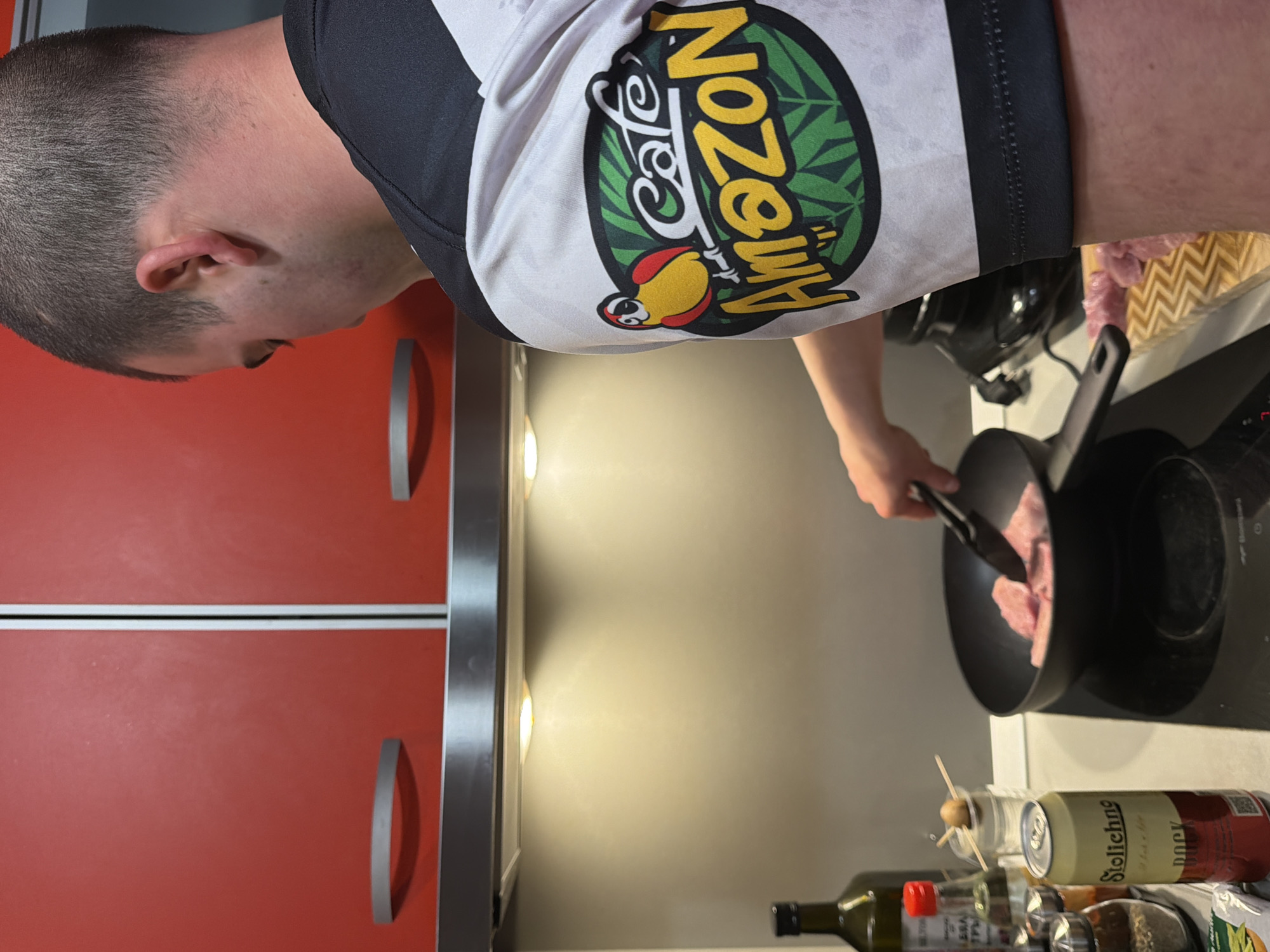An overhead view, rotated counter-clockwise, shows a person cooking in a kitchen, looking down into a frying pan. The individual has short, dark hair and is wearing a white short-sleeved shirt with black shoulders and sleeves. The prominent logo on the back of the shirt features "Cafe Amazon" in white script with a cartoon parrot (red head, yellow body, black markings) set against a green circular background with dark green leaf patterns.
The person is actively cooking, holding black tongs in their left hand to flip or stir pieces of light-colored meat (likely pork or chicken) in a black frying pan. The pan rests on a dark induction stovetop.
The kitchen setting includes red cabinets with silver bar handles on the left side of the frame. Below these cabinets, under-cabinet lighting casts a warm glow on the backsplash and countertop. On the countertop, next to the stovetop, various items are visible: a clear glass containing toothpicks, a can of "Stolichnoe" beer, a dark green glass bottle with a red cap (possibly olive oil), and another partially visible bottle. A woven basket or container filled with soft, pinkish material is also seen further back on the counter.
The overall scene suggests an indoor activity, likely meal preparation during the evening or night, given the artificial lighting. The presence of "Stolichnoe" beer is consistent with the provided location context of Varna, Bulgaria. No specific emotions are discernible from the partial view of the person.
