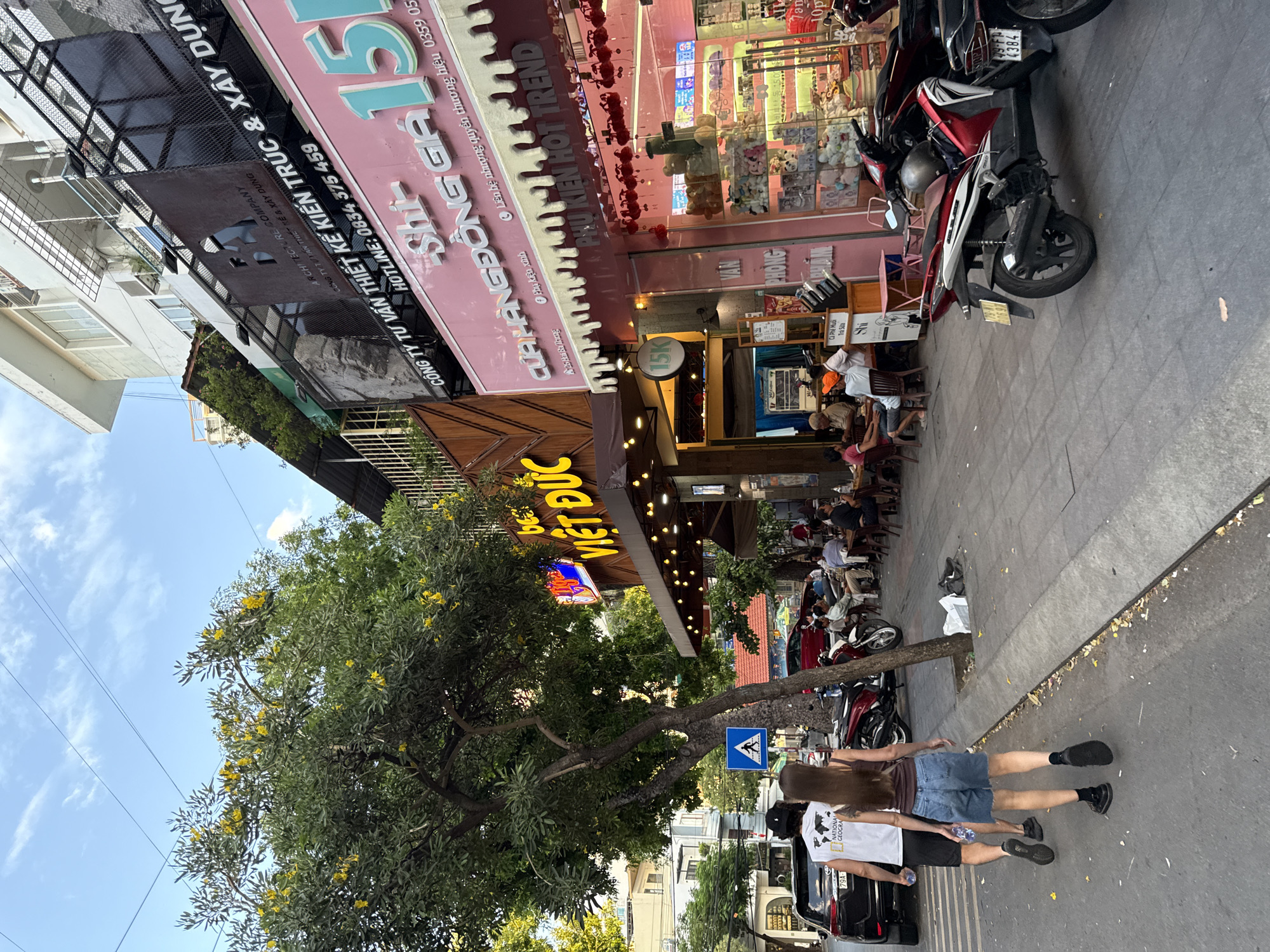 In Nha Trang, Vietnam, a street scene unfolds under a clear sky with scattered clouds. A paved sidewalk runs alongside a busy street lined with buildings housing various businesses. In the foreground, two individuals, a man and a woman, walk away from the camera, the man wearing a white t-shirt with a graphic and black shorts, and the woman in a brown tank top and denim shorts. To their left, a black SUV is parked.
A large tree with lush green foliage and yellow flowers shades part of the sidewalk. Beyond the tree, a restaurant or cafe with an outdoor seating area is visible, with several patrons seated at tables. Further down the street, storefronts display their offerings, including a bright pink establishment with signage that reads "CỬA HÀNG ĐỒNG GIA" and "PHỤ KIỆN HOT TREND." A motorbike is parked on the sidewalk in front of this store.
The architecture suggests a dense urban environment, with multi-story buildings featuring balconies and commercial spaces on the ground floor. Overhead, power lines crisscross the sky. The overall impression is one of a vibrant city street with ongoing commercial activity and pedestrian traffic.