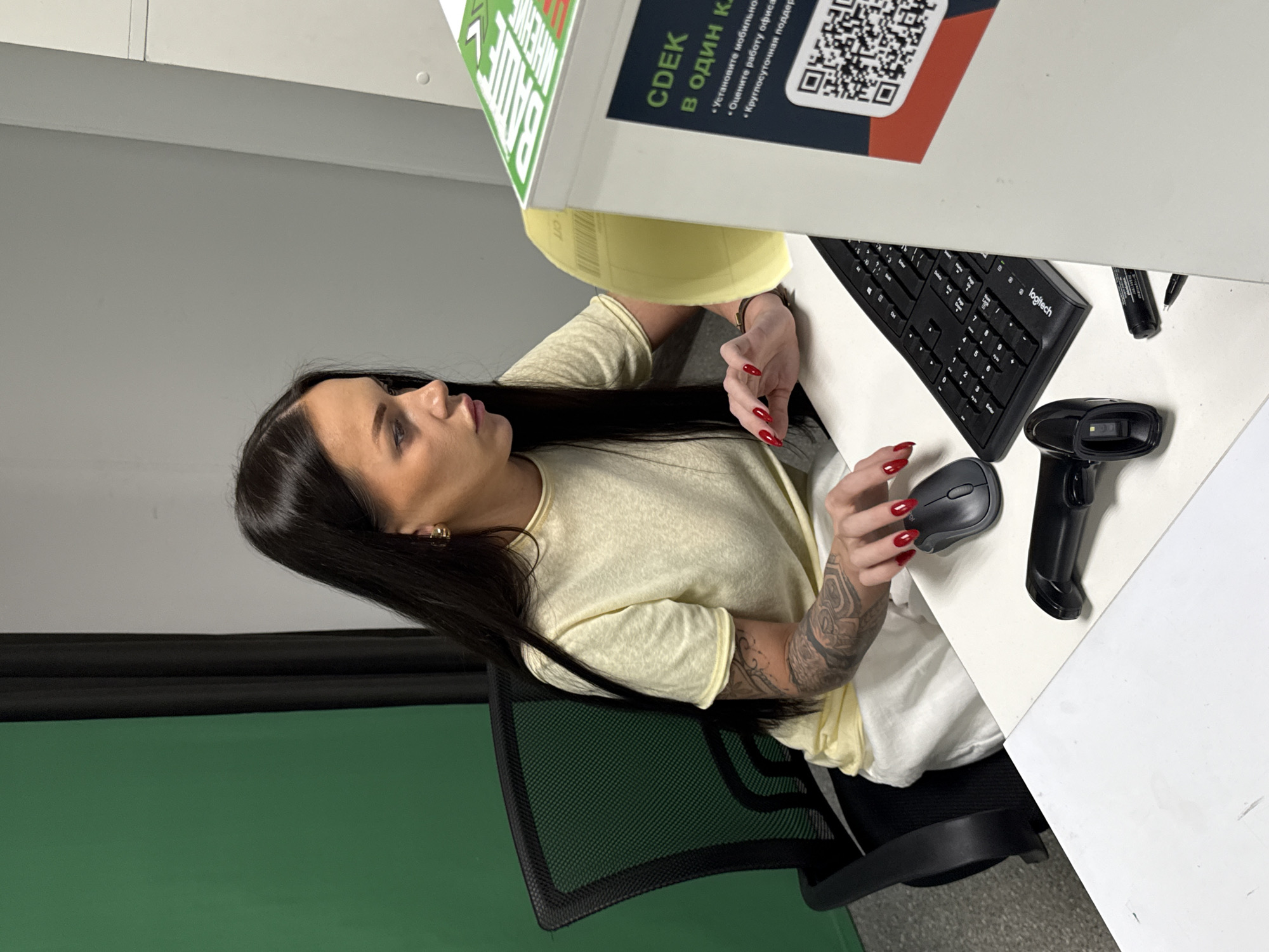 A woman with long dark hair and red nail polish is seated at a white desk, looking intently at a computer monitor. She is wearing a pale yellow t-shirt and has tattoos on her left arm. In front of her on the desk are a black computer keyboard, a grey computer mouse, and a black barcode scanner. To the right of the desk is a white cabinet with a QR code and text that appears to be in Russian. The background shows a green screen on the left and a light grey wall on the right. The scene appears to be an office environment. The location in the city of Moscow, Russia cannot be determined from the image.
