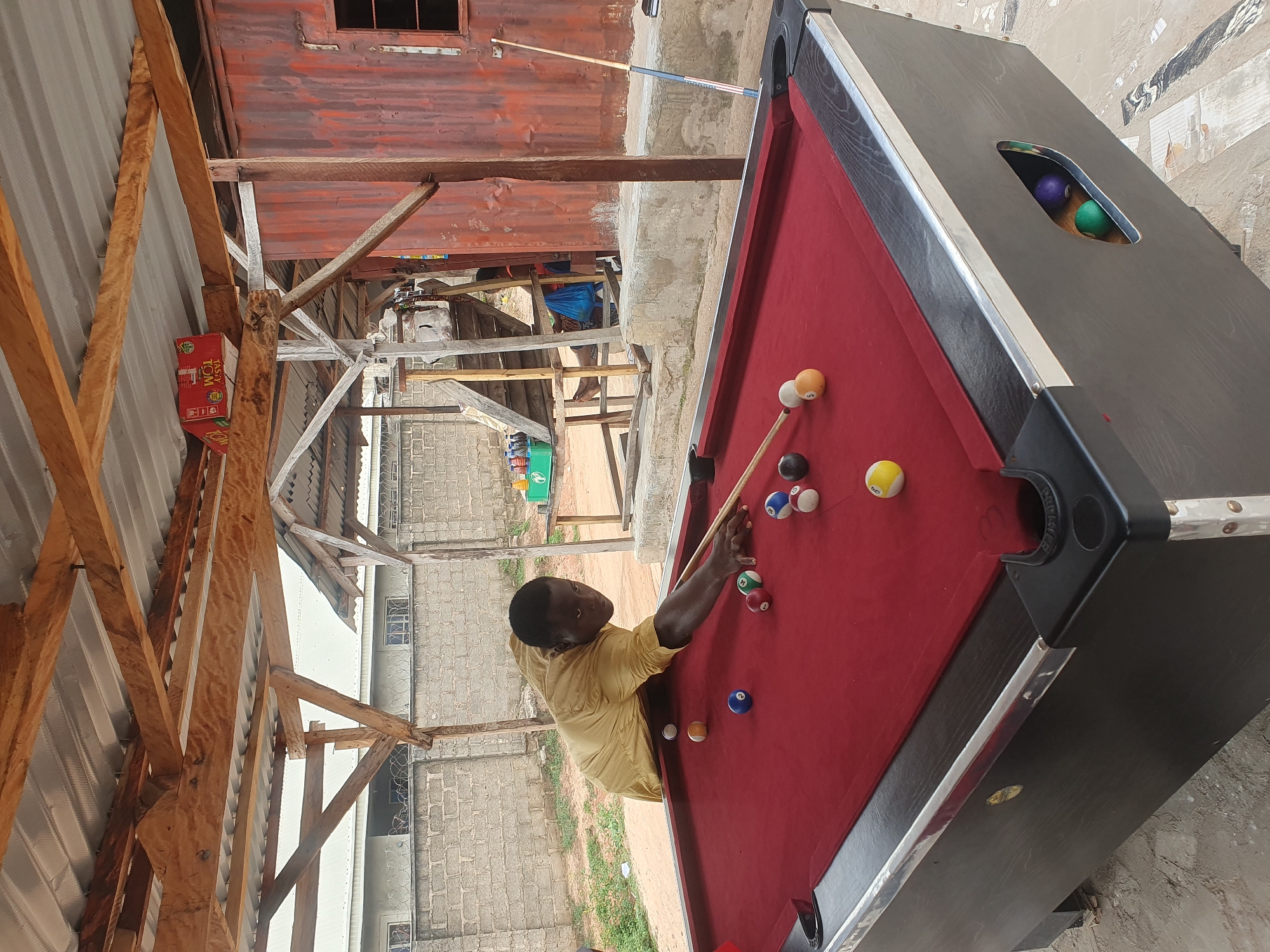 A man in a yellow shirt plays pool at an outdoor pool table. The table has a red felt surface and a black wooden frame. Several billiard balls are scattered across the table. The man is leaning over the table, holding a pool cue. The background shows a corrugated metal wall, wooden beams, and a grey brick wall. The setting appears to be an open-air establishment, possibly in Lokoja, Nigeria.