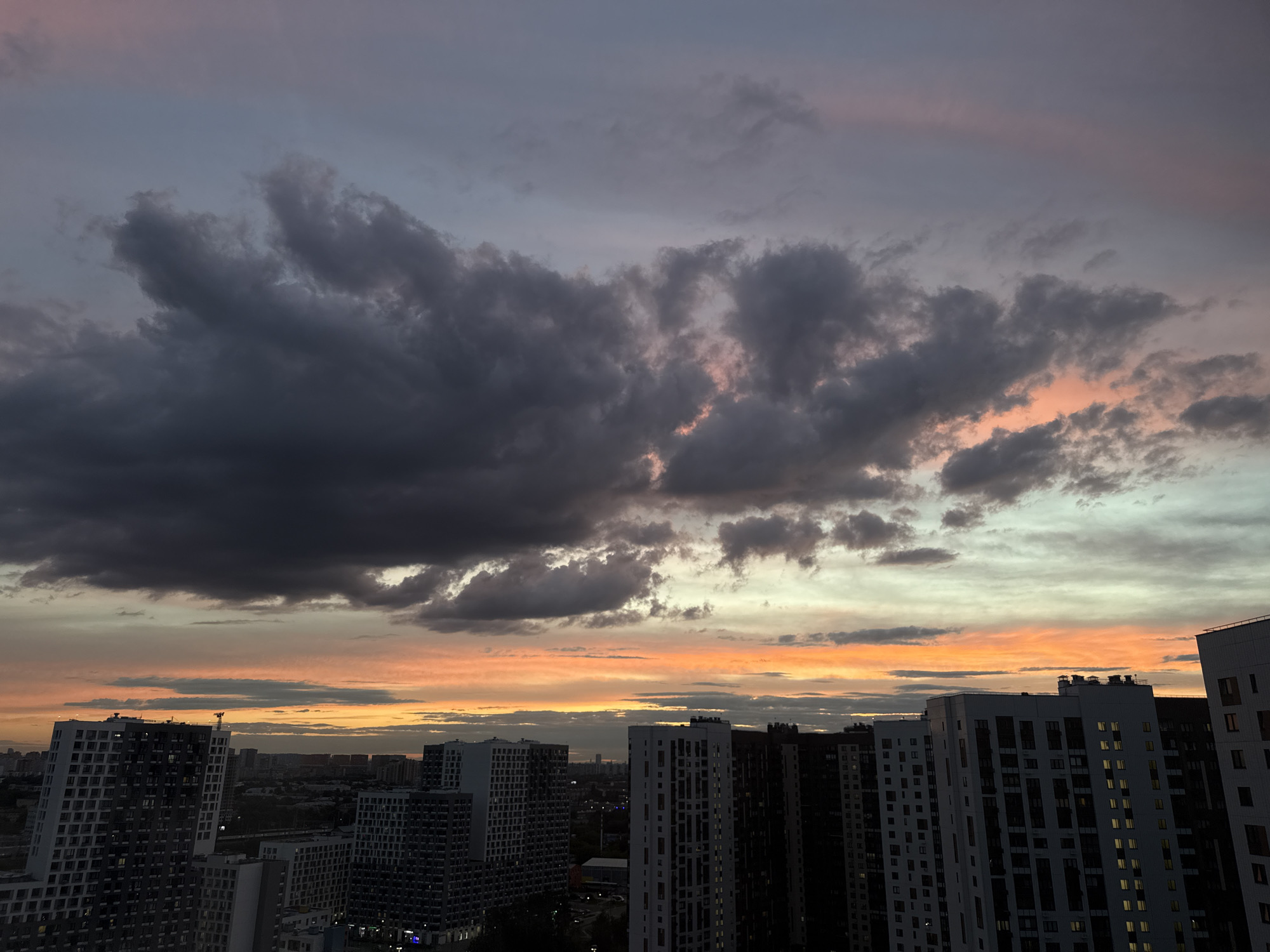This is a medium-shot, eye-level, outdoor, horizontal, color photograph taken from a high vantage point, looking out over a city at sunset. The sky is filled with dramatic clouds, ranging in color from dark gray at the top left to vibrant oranges and yellows at the horizon, with streaks of pink and pale blue also visible. Below the sky, a dense cityscape unfolds, characterized by numerous multi-story residential buildings. The buildings are dark, with many windows lit from within, suggesting it is evening. The architecture is modern, with some buildings featuring balconies and varied facade designs. The scene captures the transition from day to night in an urban environment. The location is Lyubertsy, Russia.