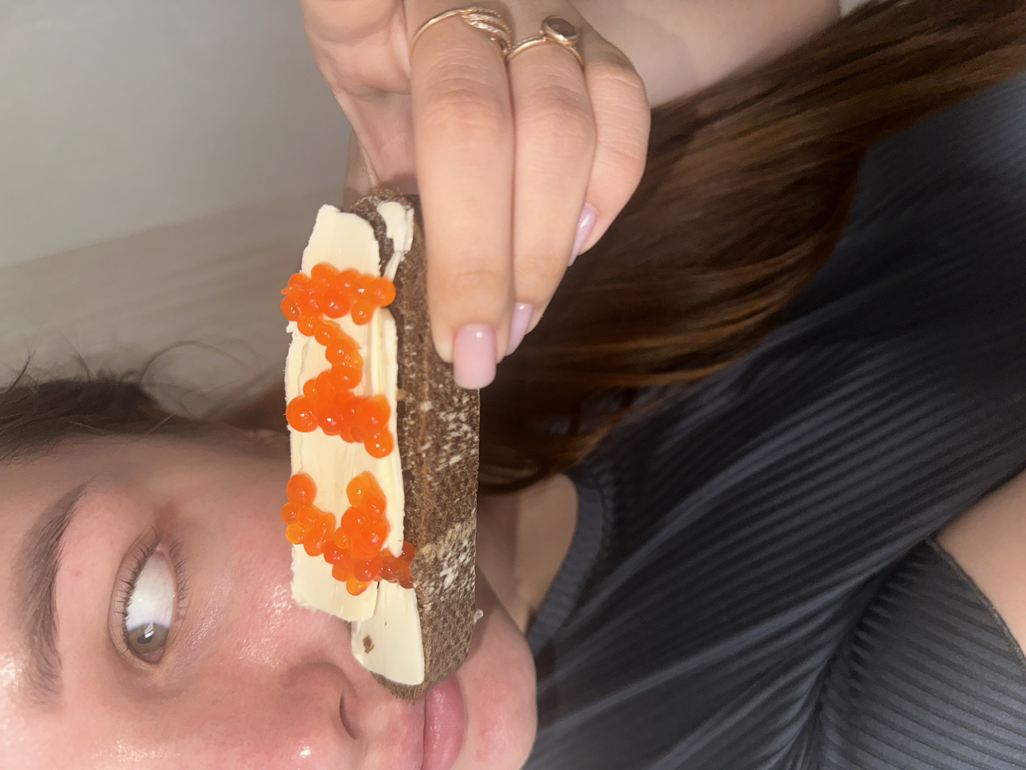 A person is holding a slice of dark bread with butter and red caviar. The person has brown hair and pink nail polish. They are wearing two rings on their fingers. The shirt they are wearing is black and grey striped. The location is Chaikovsky, Russia.