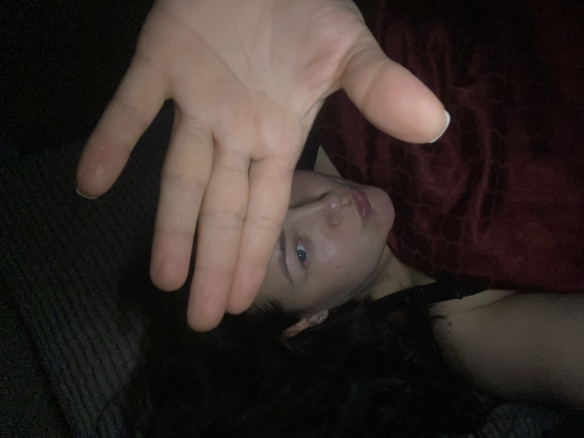 A woman is lying down, partially obscured by a large hand held up towards the camera. The woman has dark hair, fair skin, and blue eyes. She is wearing a black top. A maroon colored fabric is draped over part of her upper body. The background appears dark, with a section of gray fabric visible to the left. The location is unknown; there is no indication of Sochi, Russia, or any specific city.