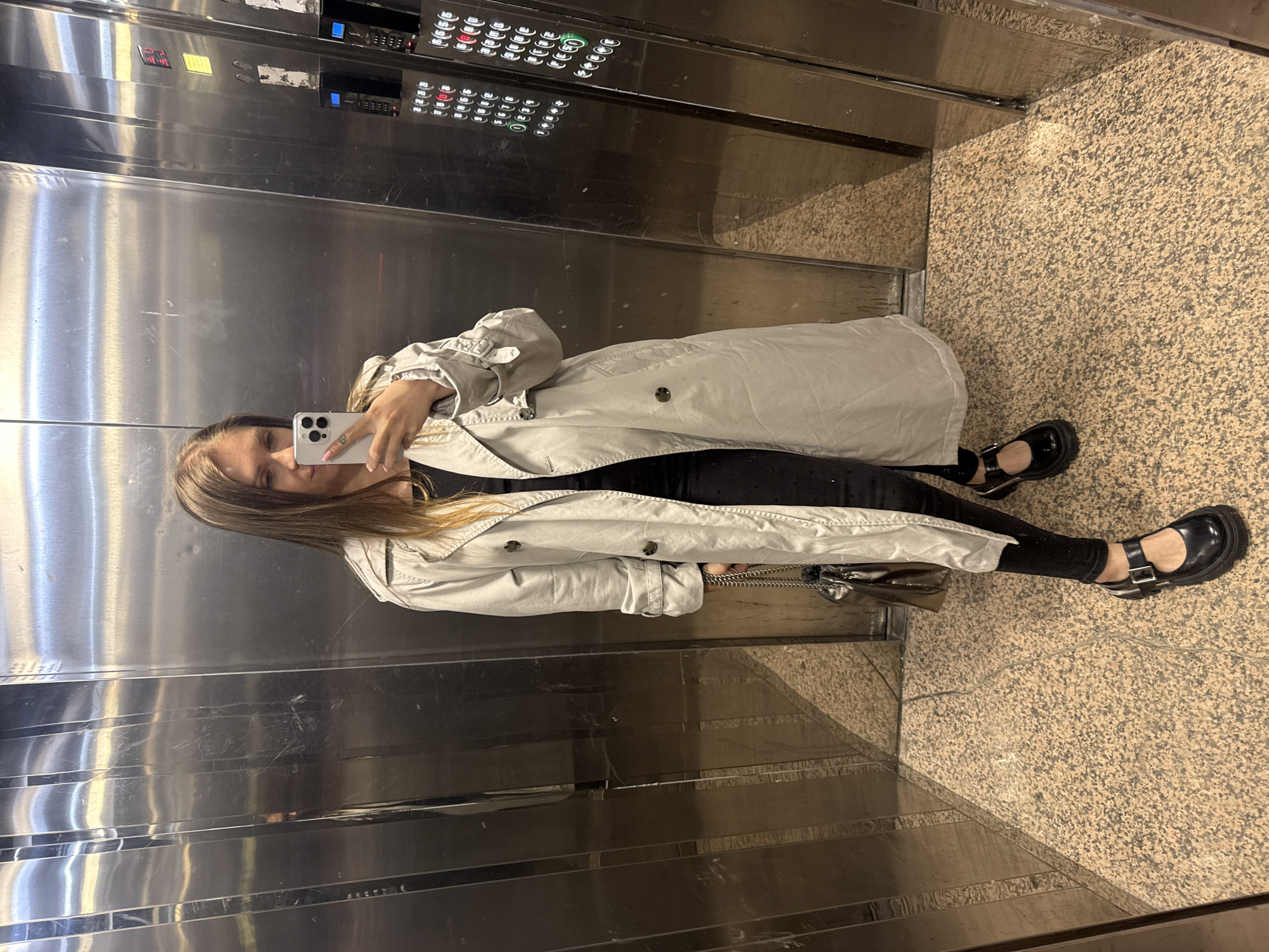 The image shows a woman in an elevator, seemingly taking a selfie in the reflective surface. The elevator has a stainless-steel interior and a floor with a speckled pattern. The elevator panel with floor buttons is visible. The woman is wearing a light-colored trench coat, black leggings, and black Mary Jane shoes. She is holding a smartphone. The location is not apparent from the image. It is not possible to determine if this image was taken in Batumi, Georgia.